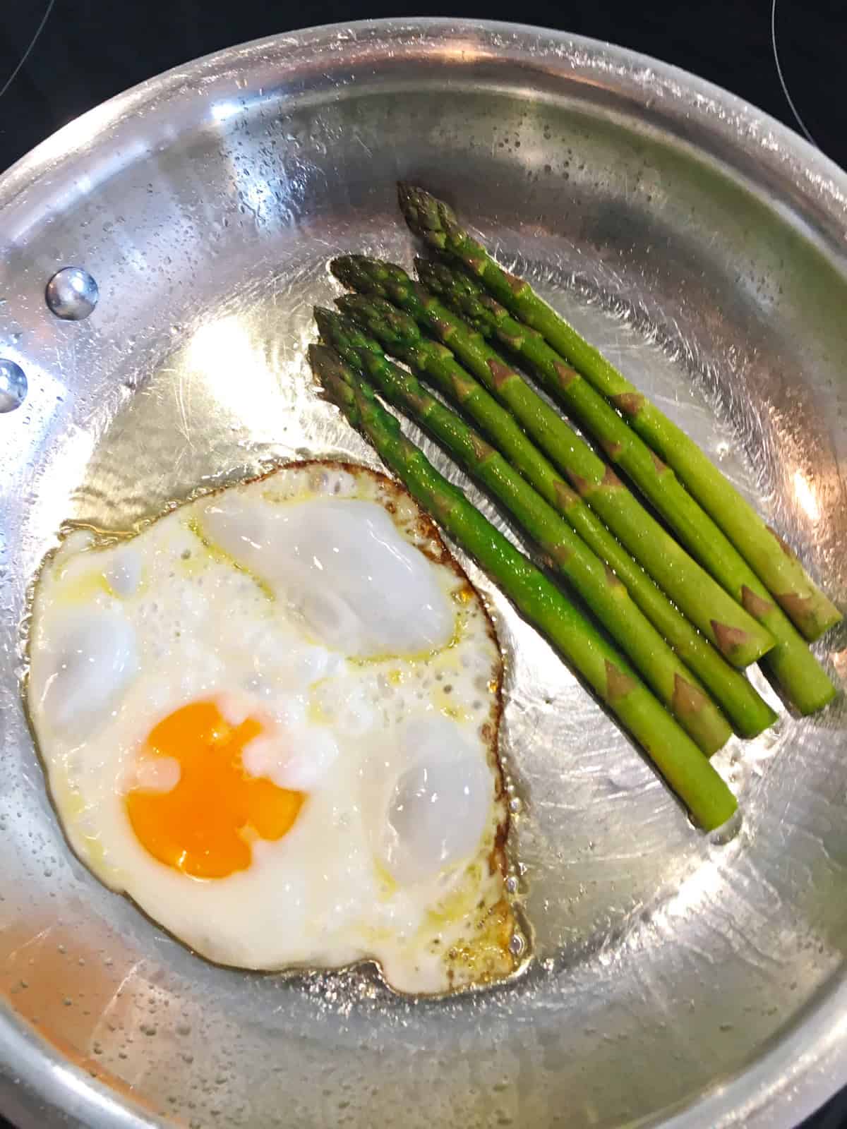 A sunny side up egg and six asparagus spears frying in butter in a stainless steel skillet.