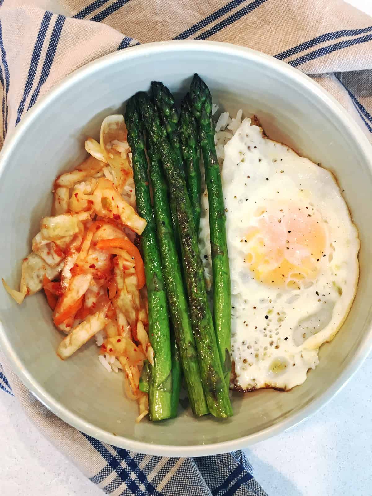 Kimchi breakfast bowl with asparagus and eggs in an off-white ceramic bowl with a blue and tan patterned napkin