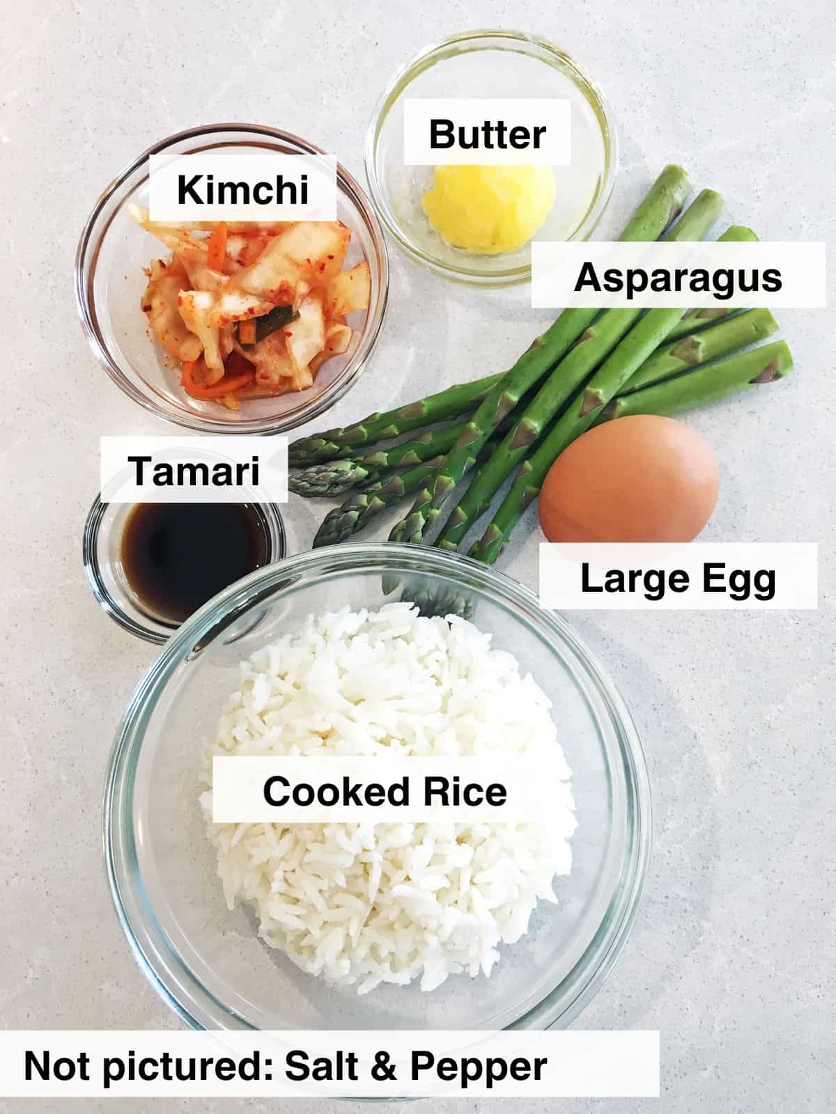 White rice, kimchi, butter and tamari in glass food prep bowls alongside a whole brown egg and six stalks of trimmed asparagus.