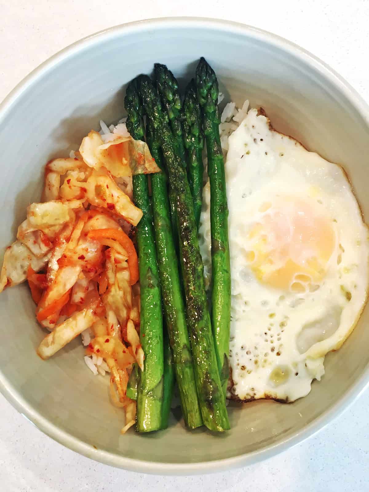 Kimchi, fried egg and asparagus spears over white rice in an off-white bowl atop a light grey marble countertop