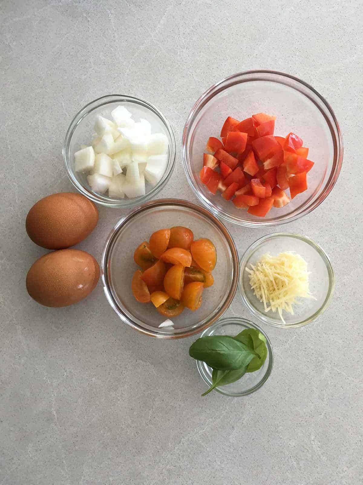 Two whole brown eggs alongside small glass food prep bowls containing chopped onions, red peppers, cherry tomatoes, shredded Parmesan cheese and fresh basil leaves