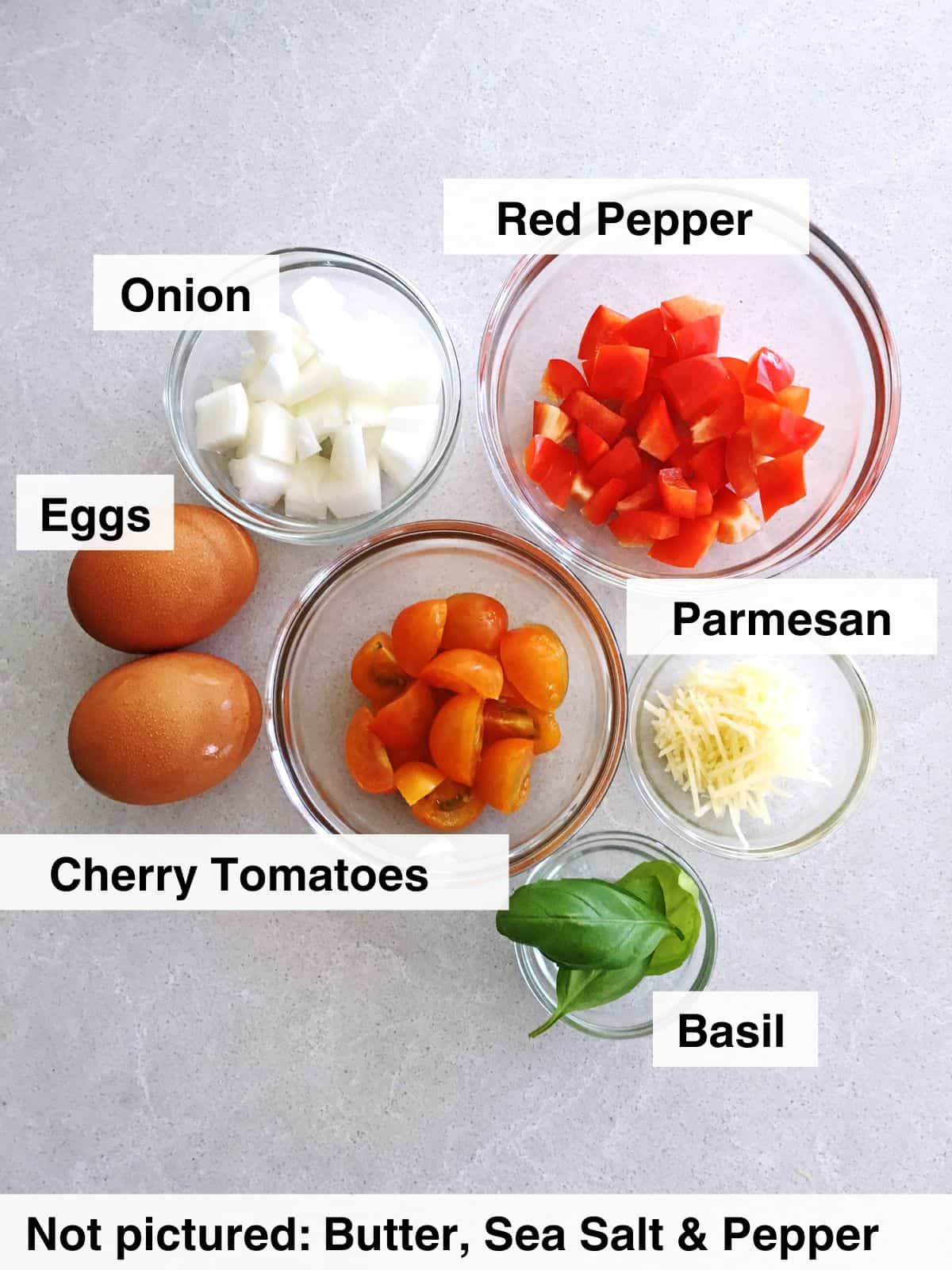 Two whole brown eggs alongside small glass food prep bowls containing chopped onions, red peppers, cherry tomatoes, shredded Parmesan cheese and fresh basil leaves