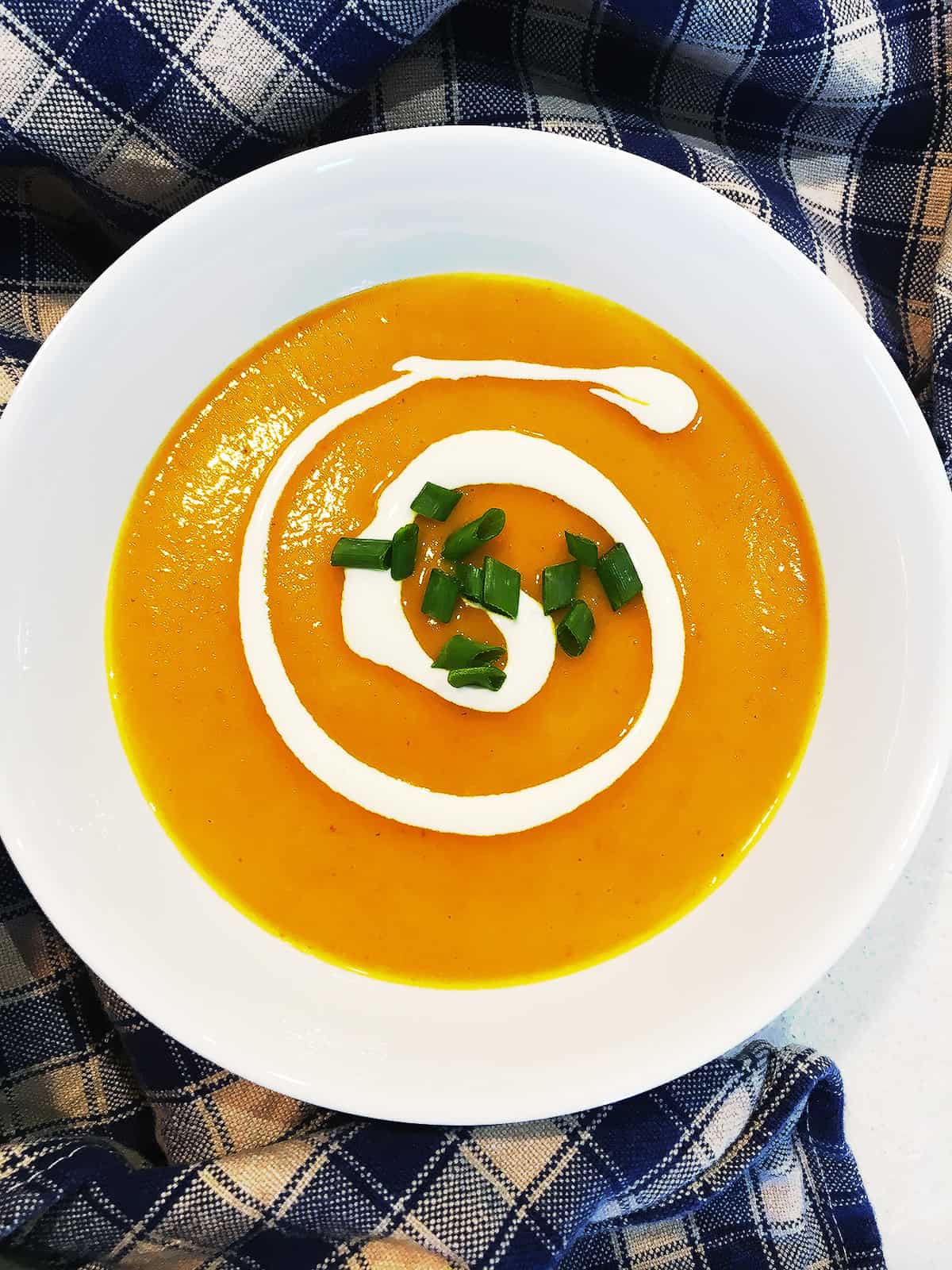 Creamy carrot soup with potatoes garnished with sour cream and chives and adorned with a navy blue and tan plaid napkin.