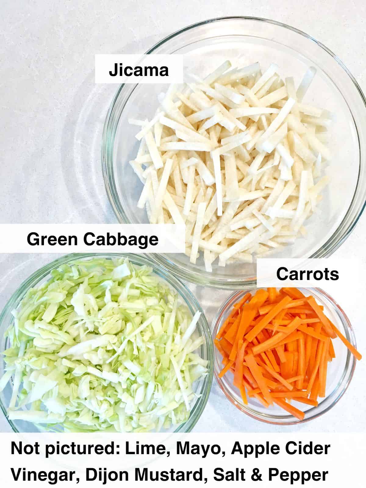 Sliced green cabbage, jicama and carrots in three glass bowls atop a light grey marble countertop.