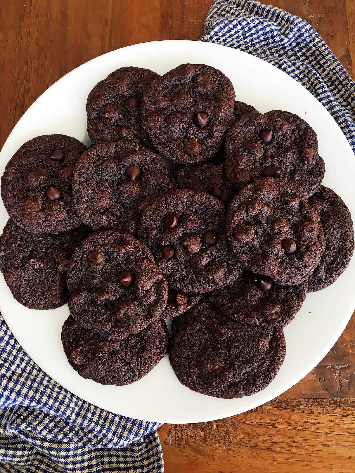 Freshly baked soft and chewy double chocolate chip cookies on a white serving plate atop a wood table.