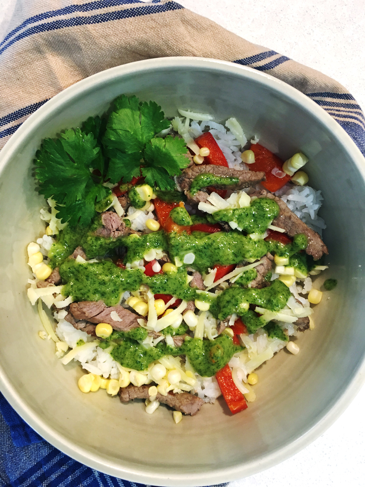 Veggie and steak rice bowl with cilantro lime pesto in a tan ceramic bowl with a blue and tan striped napkin