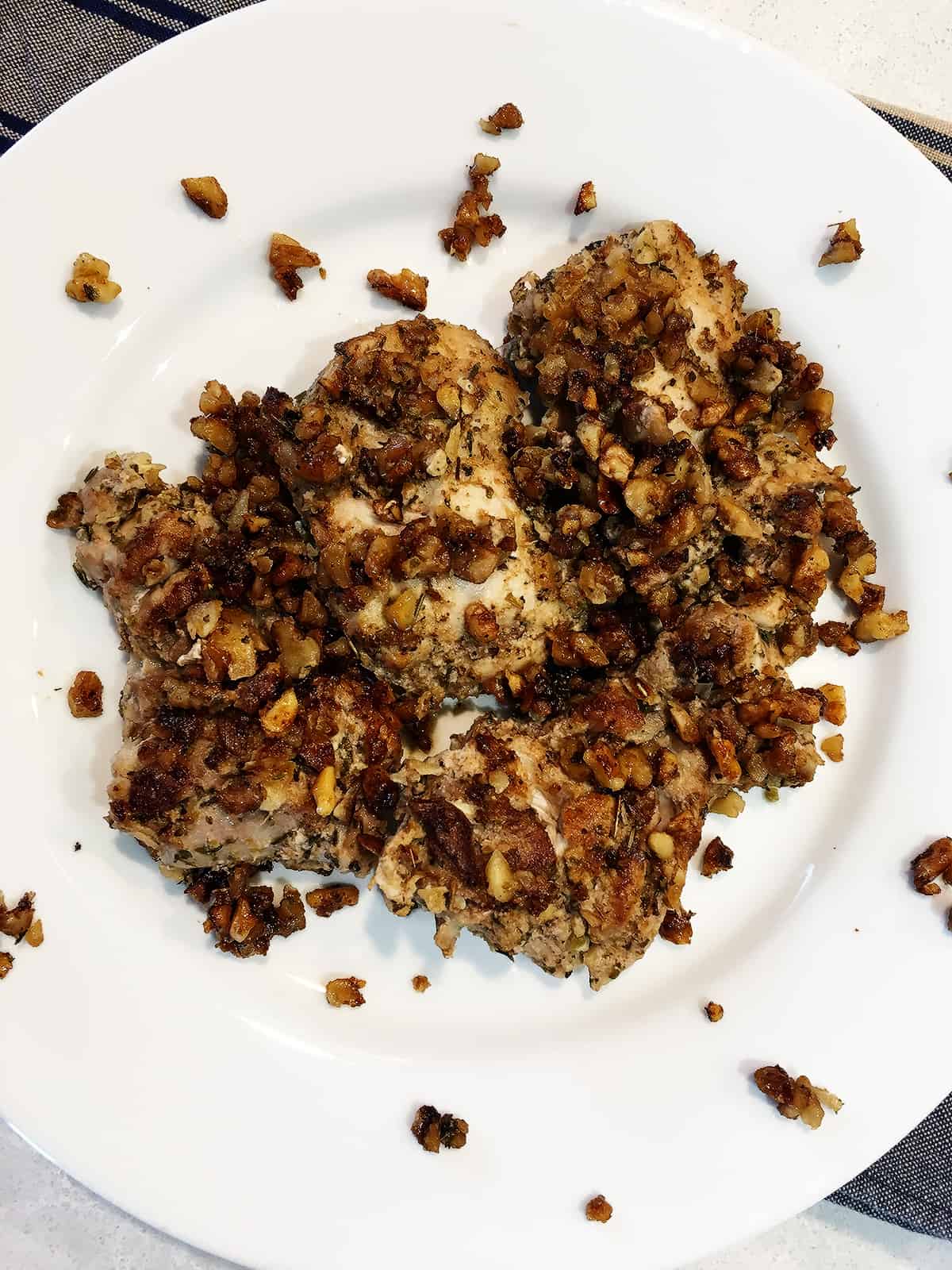 Herb and walnut crusted chicken thighs on a white plate atop a tan and navy napkin.