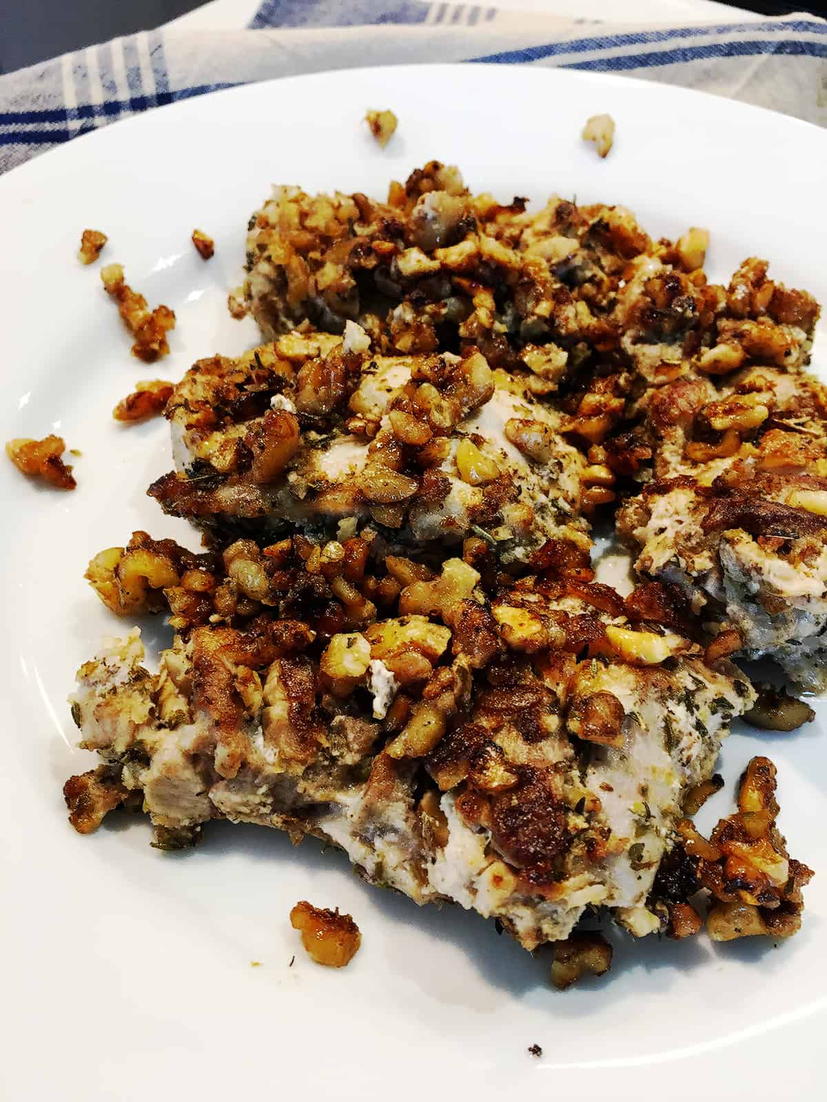 Close up of walnut and herb crusted chicken thighs on a white serving plate with a blue and tan plaid napkin.