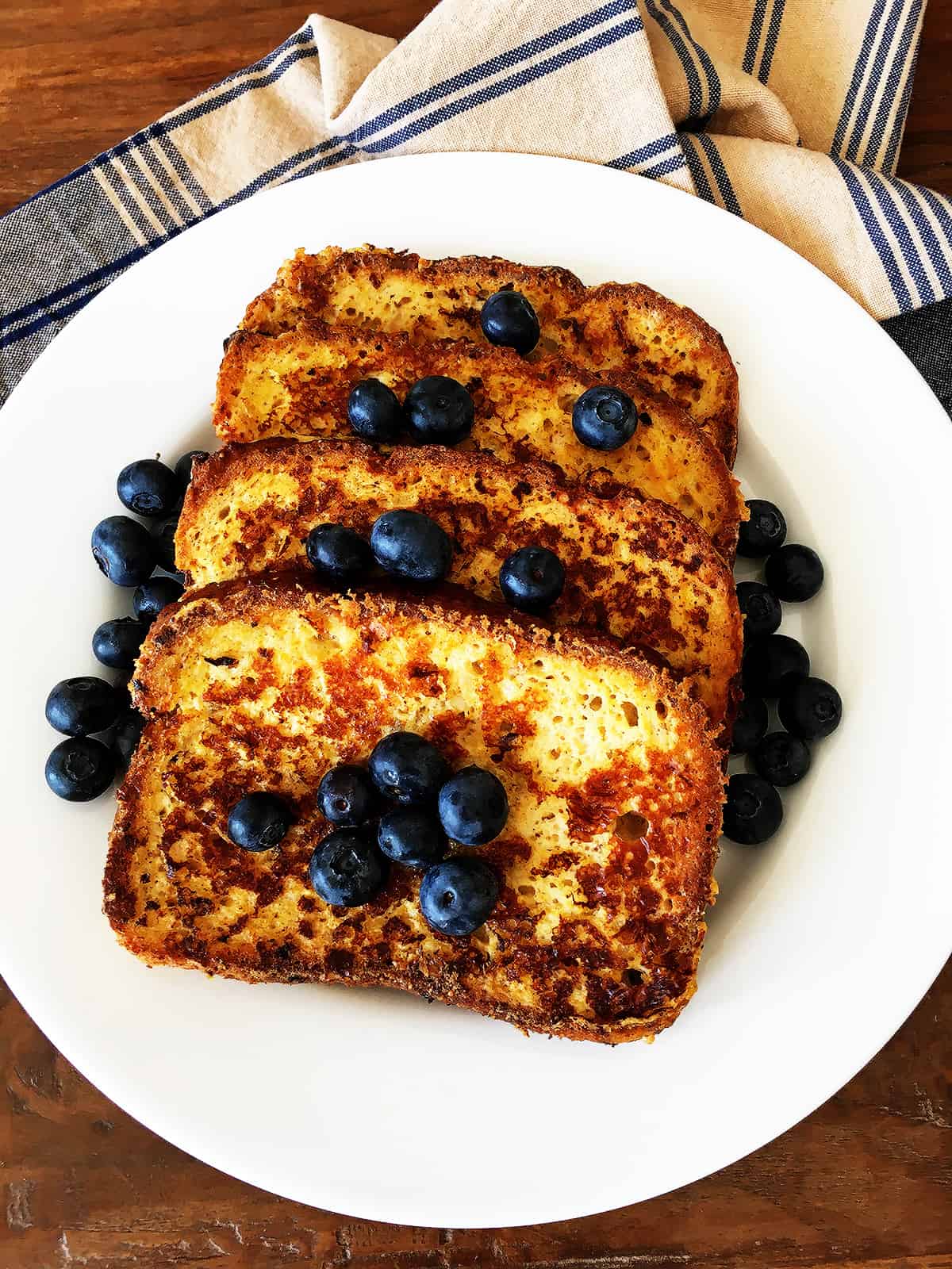 French toast with fresh blueberries on a white plate with a blue and tan plaid napkin