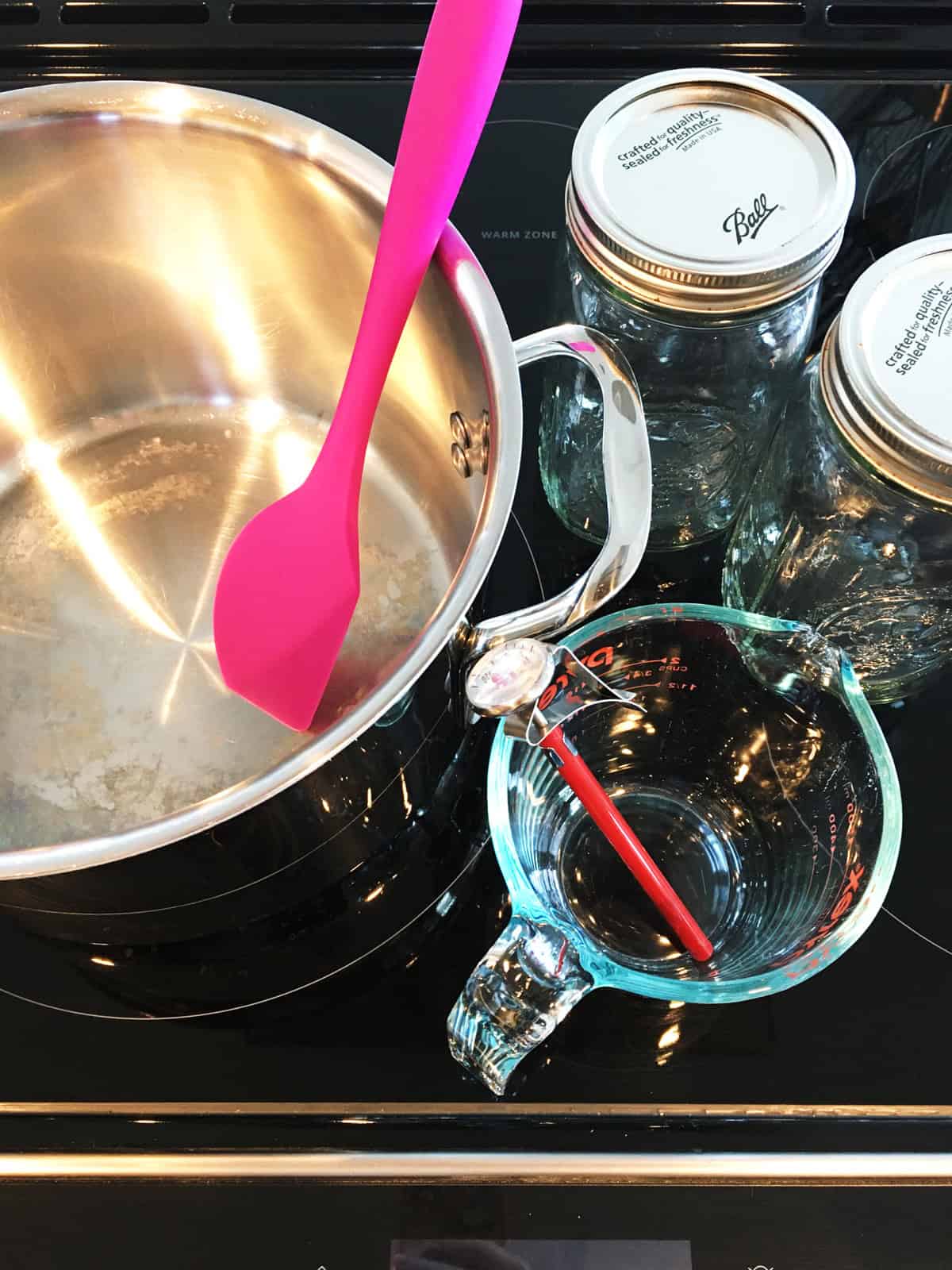 Stainless steel soup pot, spatula, mason jars, measuring cup and thermometer on a black stove top