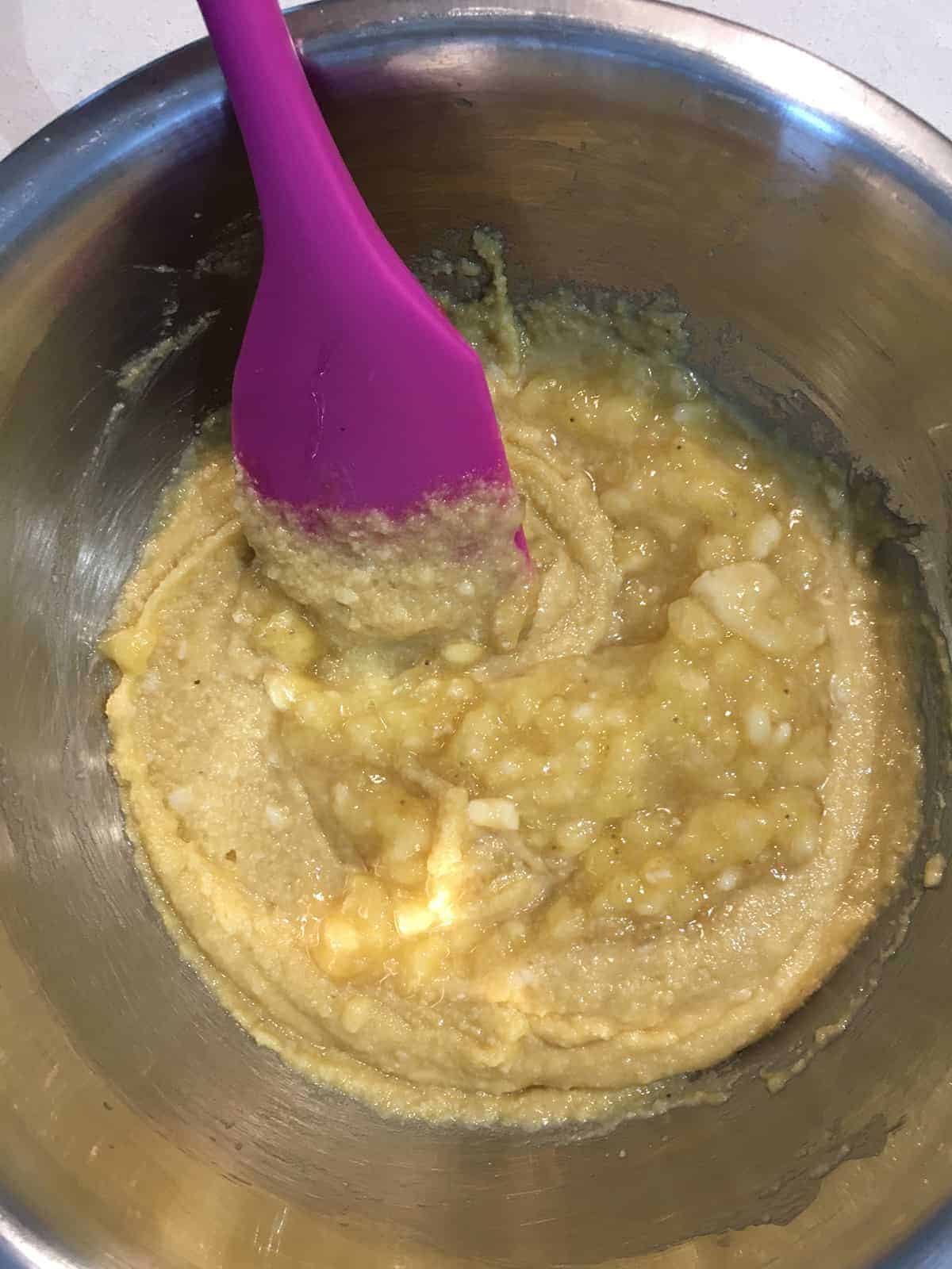 Banana bread batter in a stainless steel mixing bowl with a purple spatula