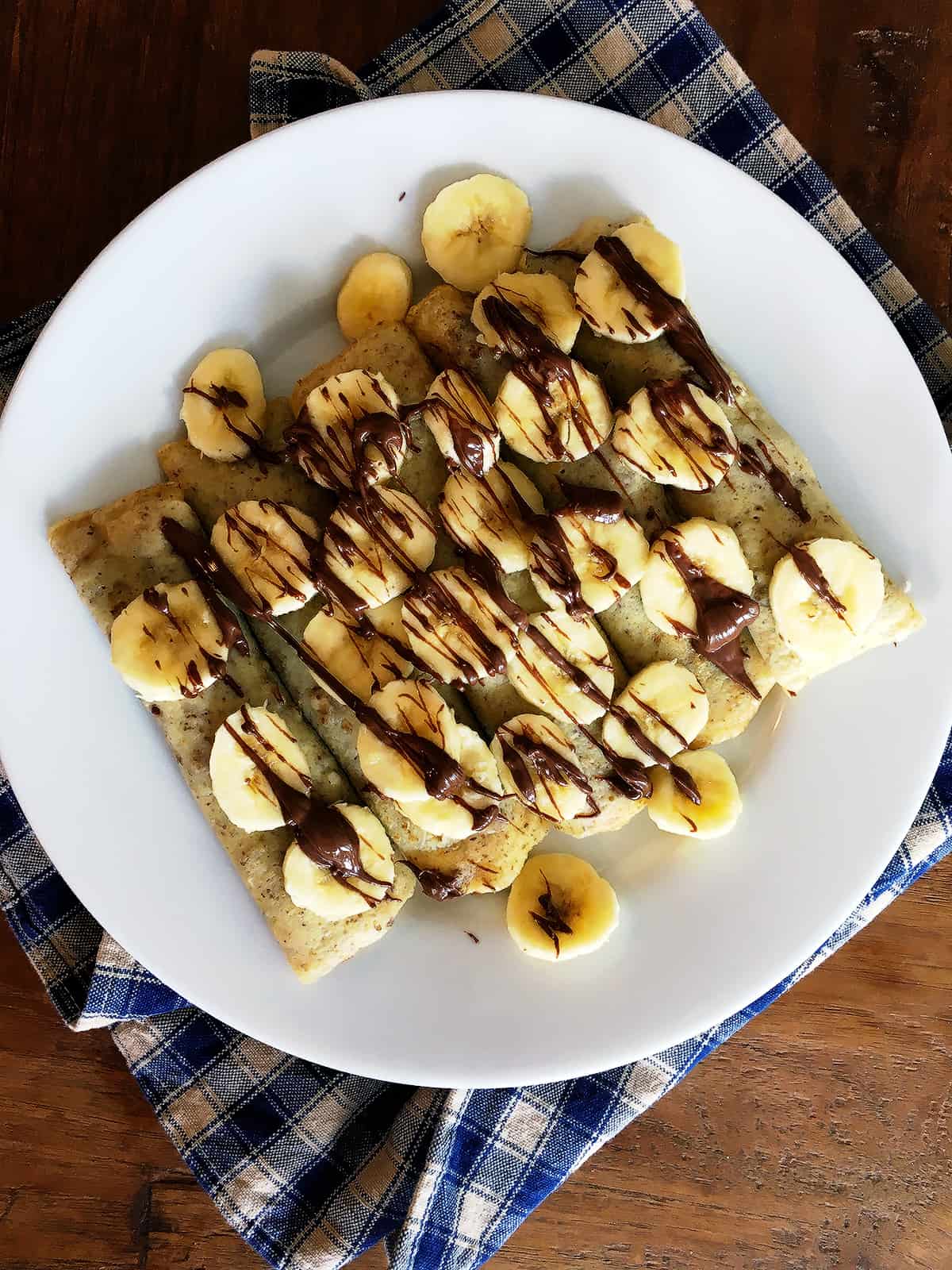 Banana Nutella crepes on a white plate with a tan and blue plaid napkin.