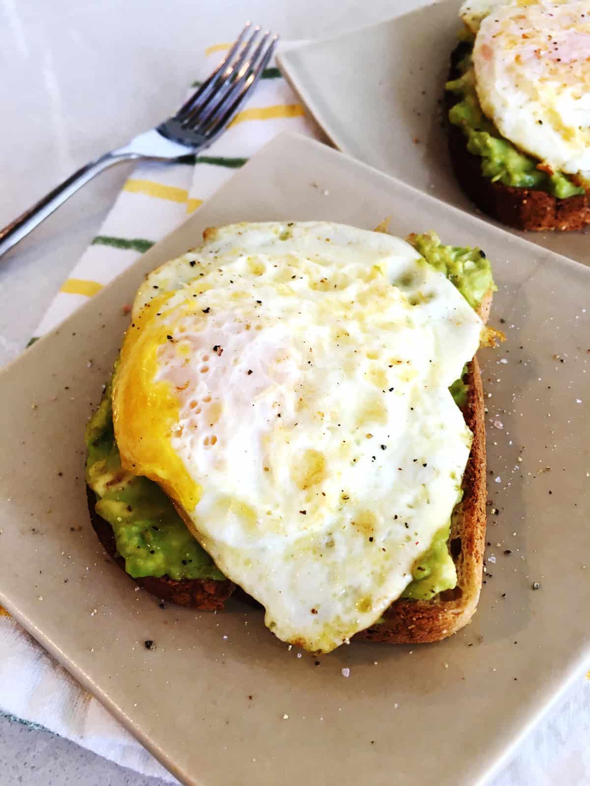 Avocado toast with egg on tan ceramic plates atop a light grey marble countertop.