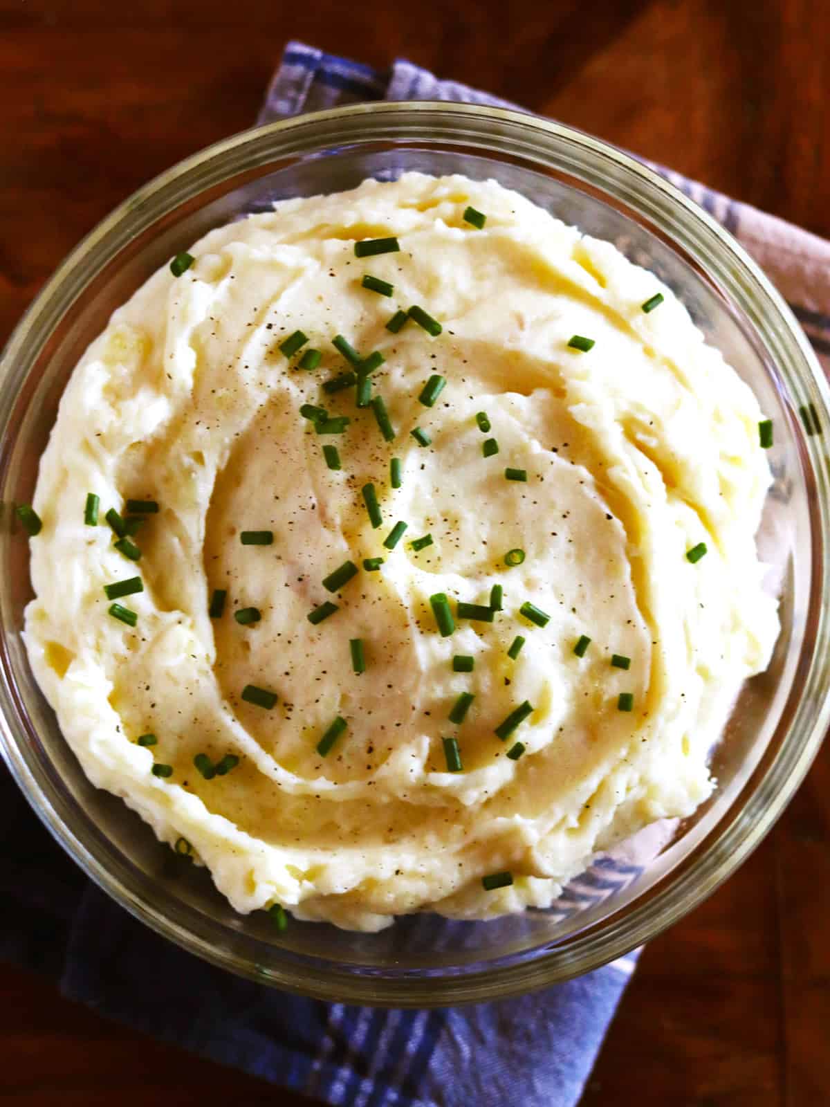 Homemade mashed potatoes garnished with fresh chives in a glass bowl with a blue plaid napkin.