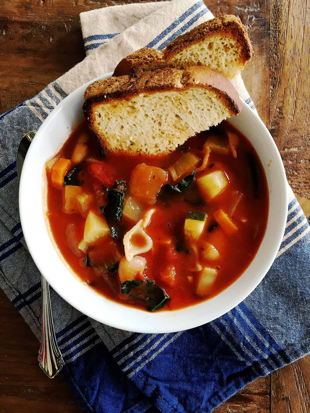 Minestrone soup and toasted bread in a white bowl atop a brown wood table.