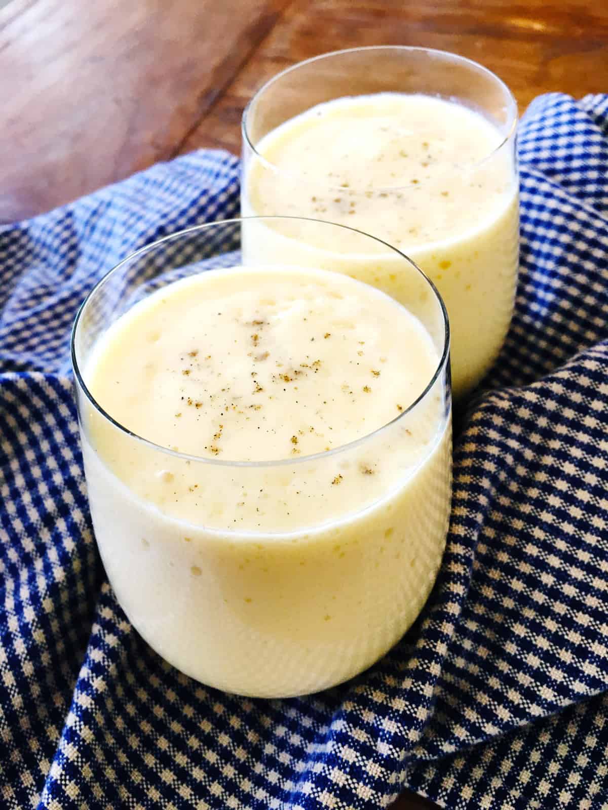 Two mango lassi drinks topped with cardamom on a wood table with a blue checkered napkin.