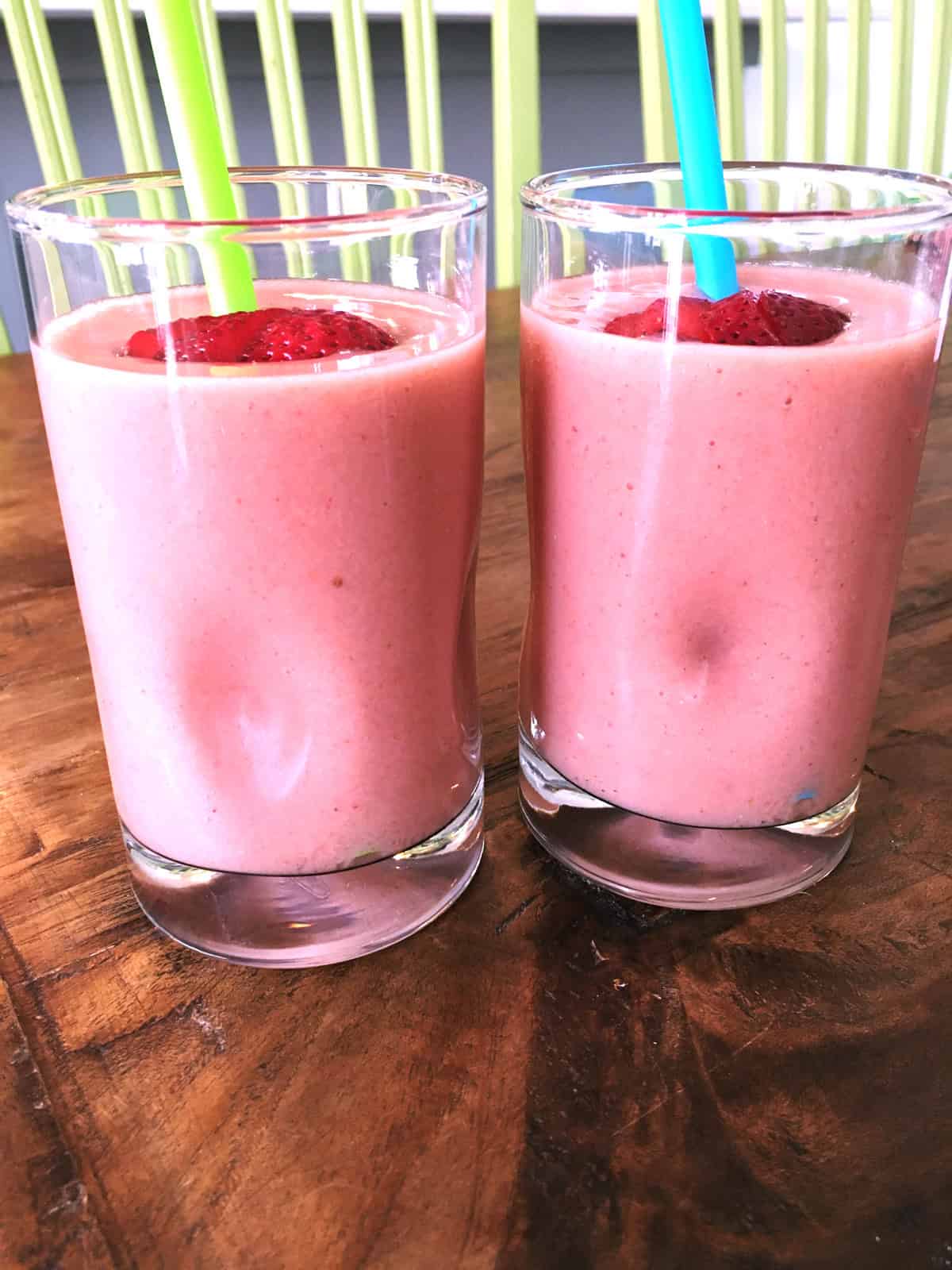 Two strawberry banana smoothies with green and blue straws atop a brown wood table.