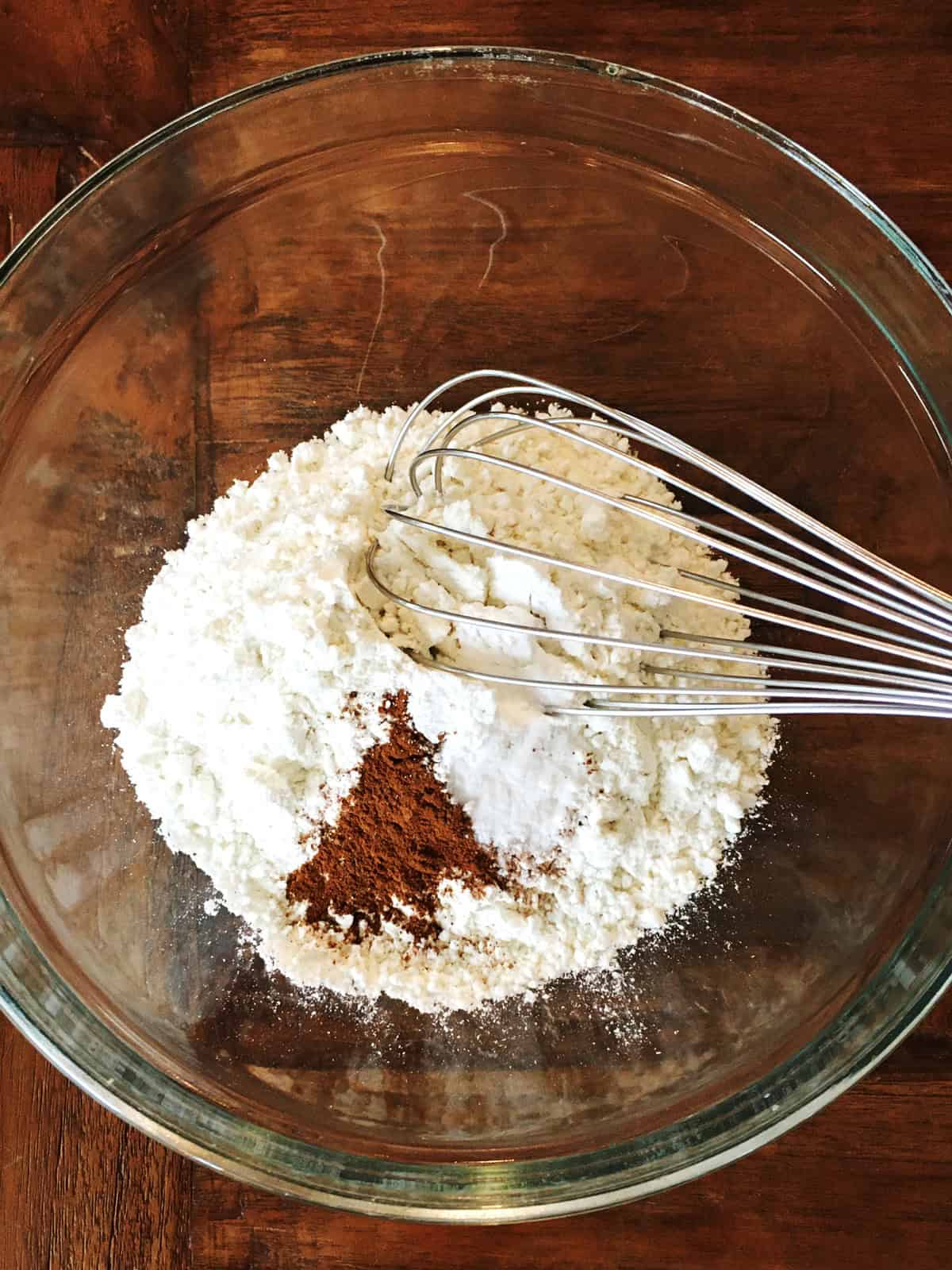 Flour, baking powder, baking soda, cinnamon and salt with a silver whisk in a glass mixing bowl.