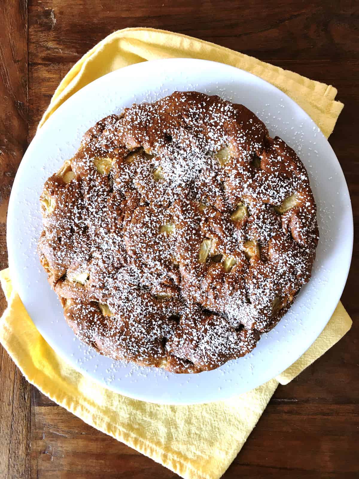 Apple cake dusted with powdered sugar on a white plate atop a brown wood table.