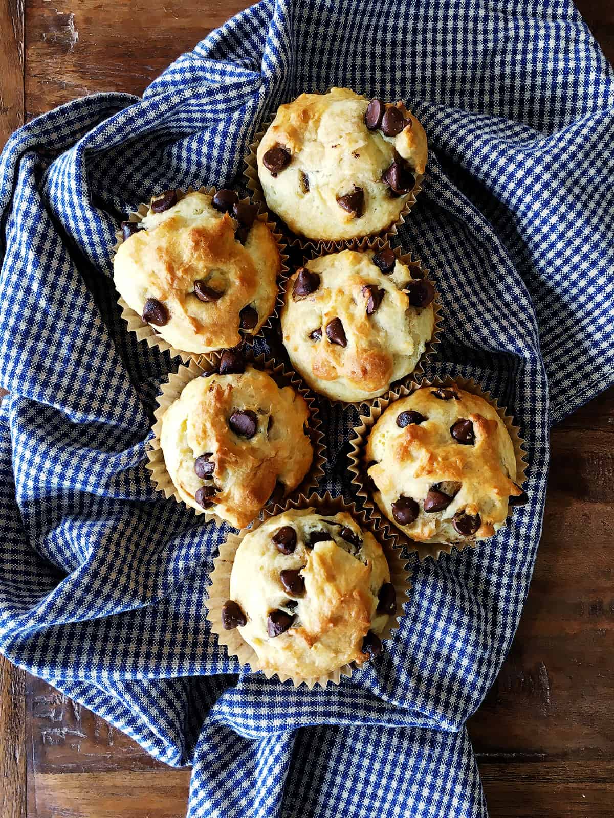 Chocolate chip muffins on a blue and tan plaid napkin atop a brown wood table.