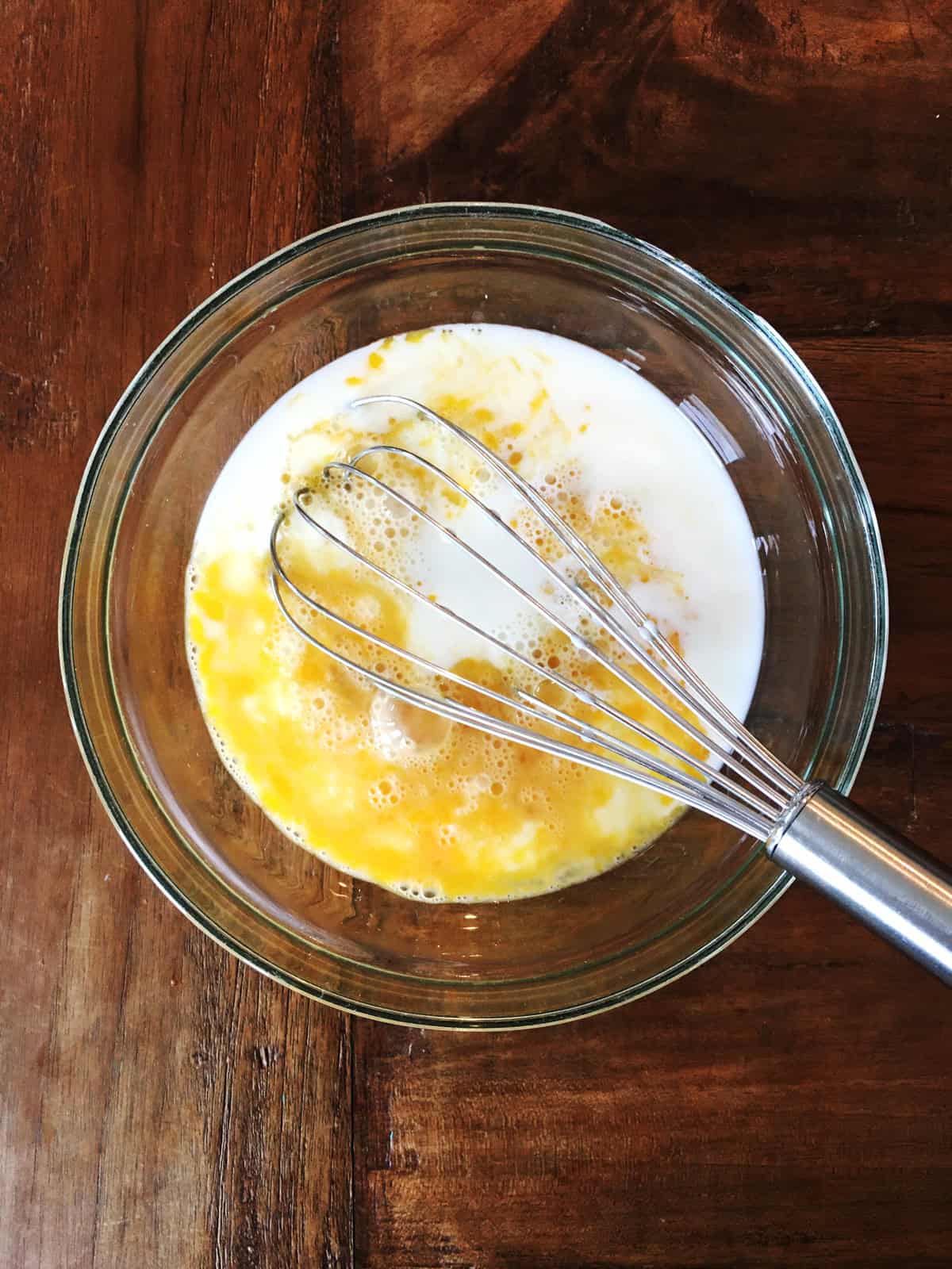 Wet ingredients for pancakes in a glass mixing bowl with a stainless steel whisk