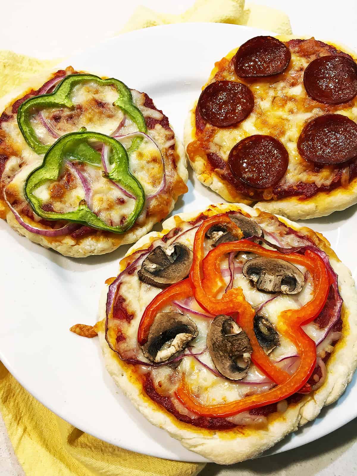Three mini pizzas with various toppings on a white plate with a blue and tan plaid napkin