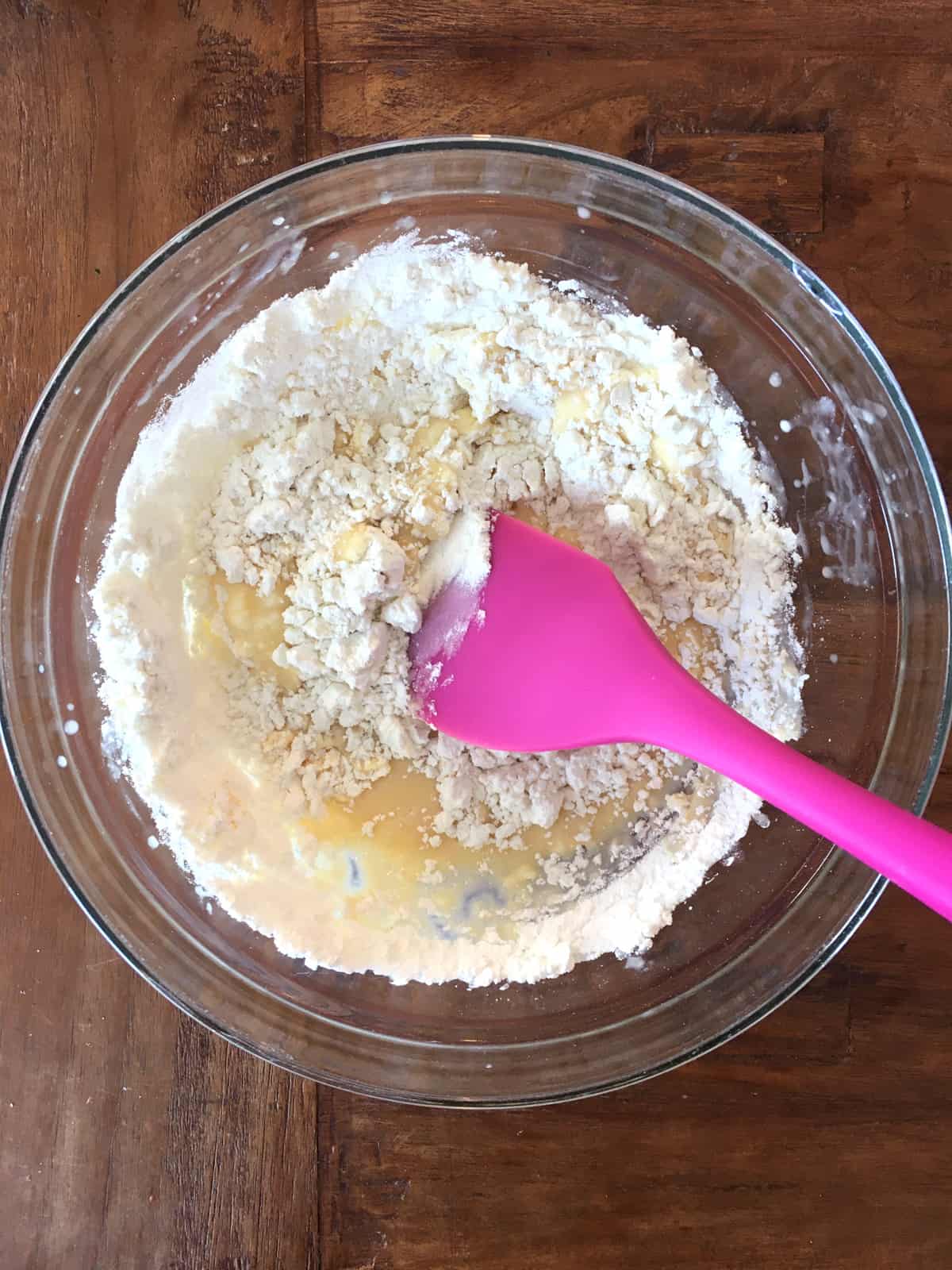 Folding dry ingredients into wet in a glass bowl with a purple silicone spatula.