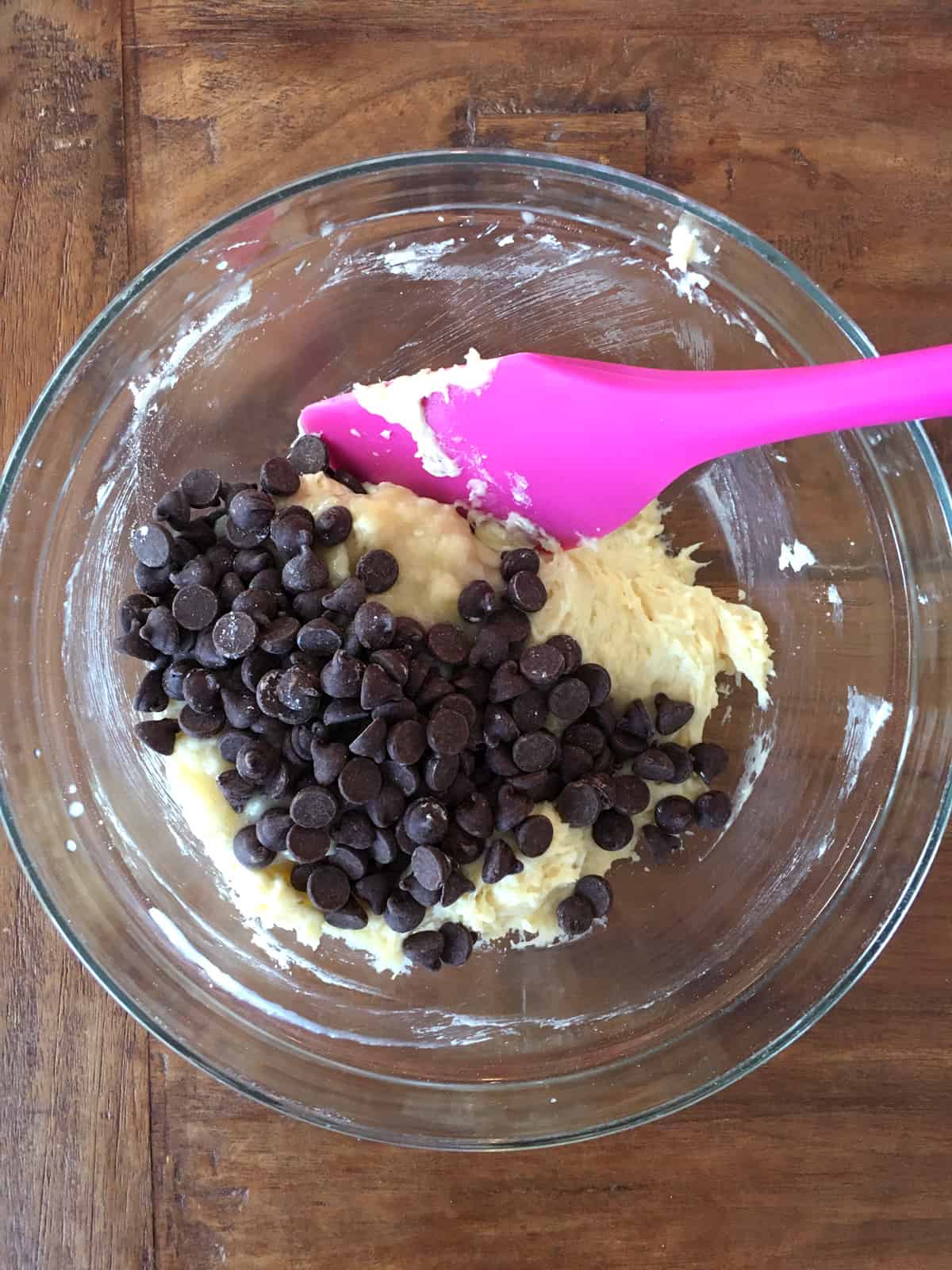 Folding bananas and chocolate chips into muffin batter with a purple silicone spatula.