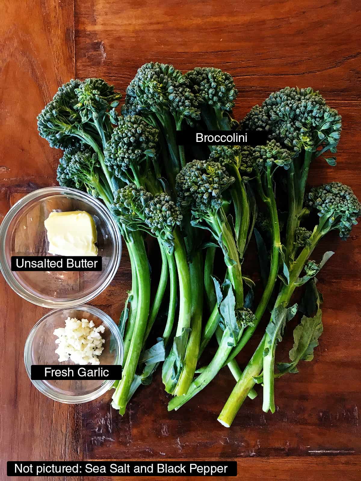 Fresh broccolini stalks next to small glass bowls of butter and minced garlic on a brown wood table.