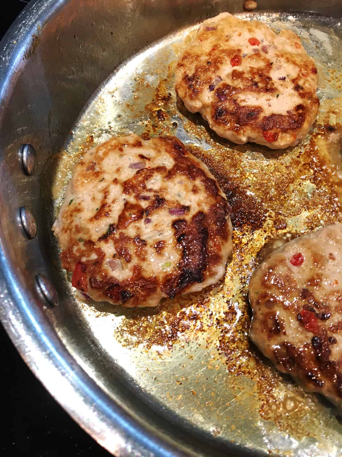 Turkey burgers pan frying in olive oil in a stainless steel skillet.