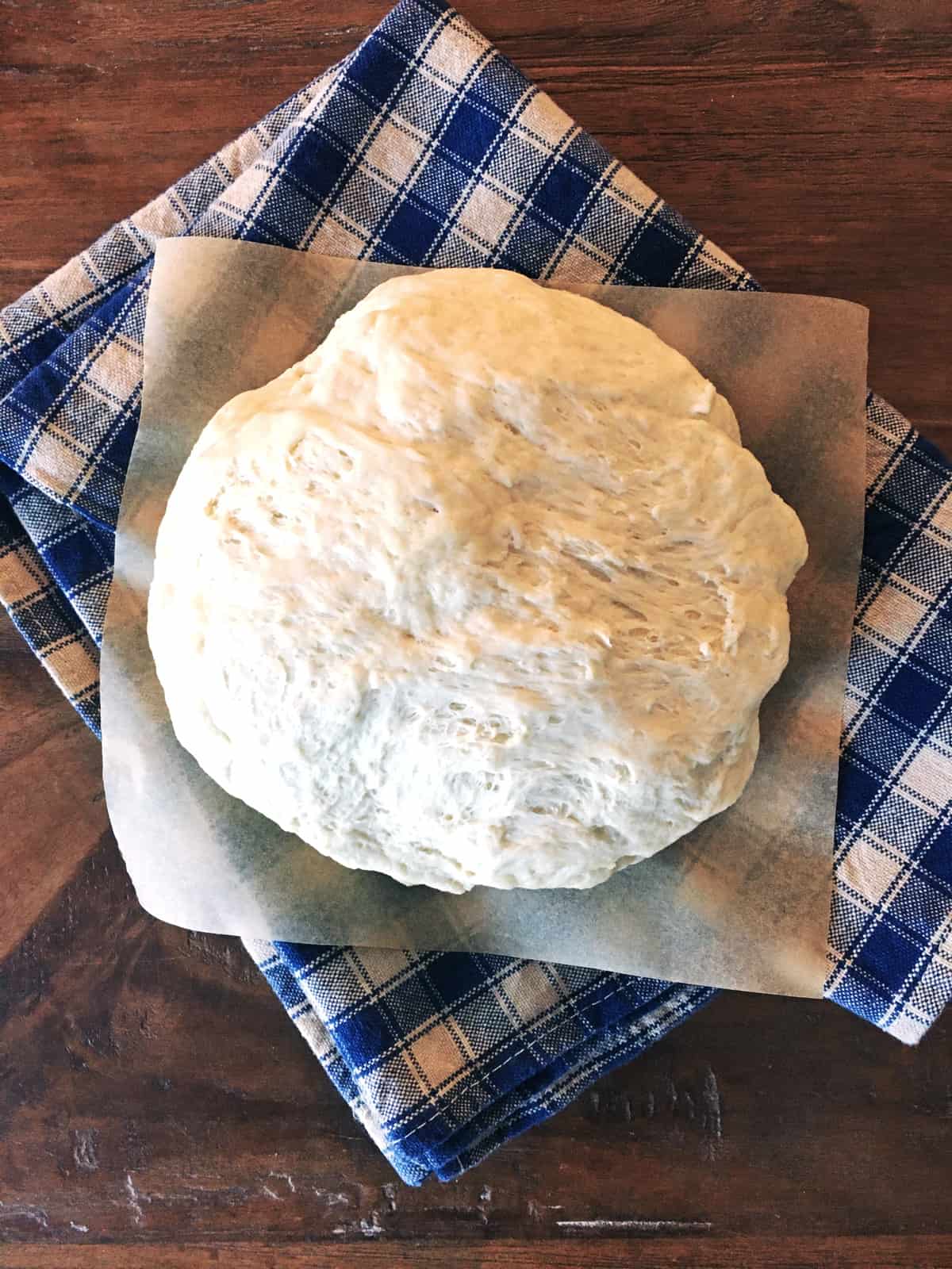 Homemade pizza dough on parchment paper with a blue and tan plaid napkin.