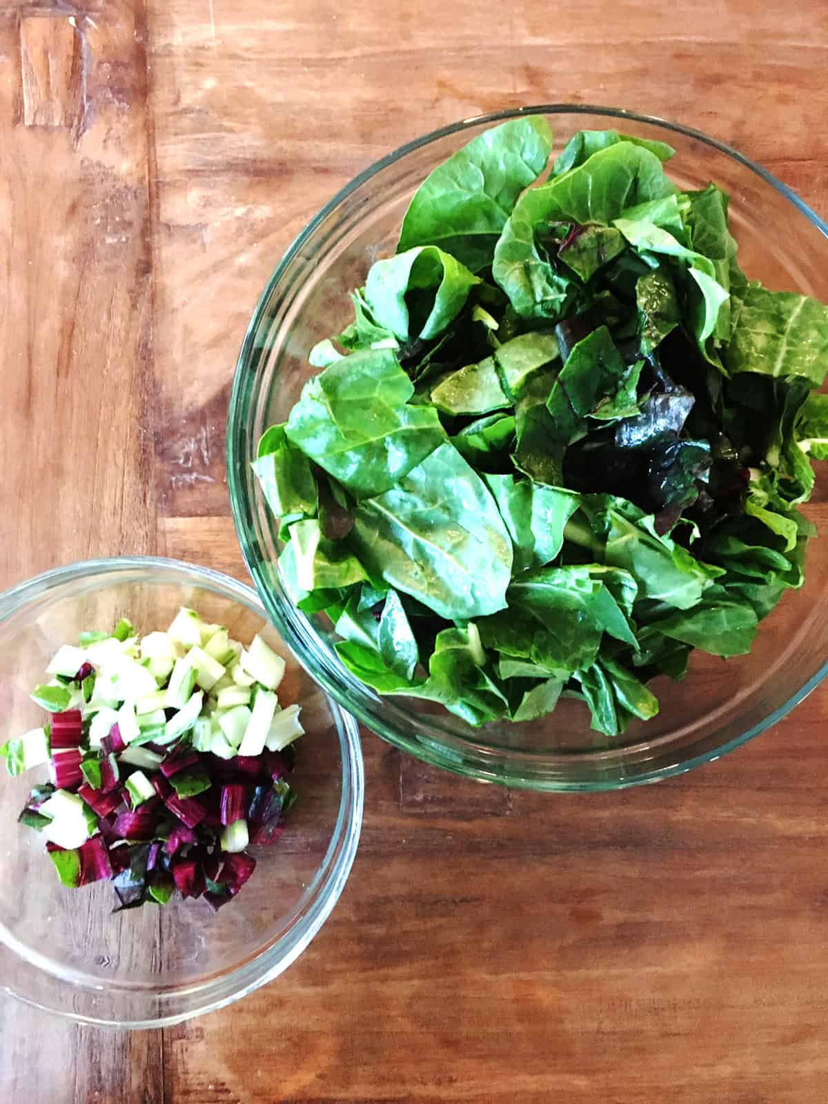 Swiss chard leaves and stems in two glass food prep bowls on a brown wood table.