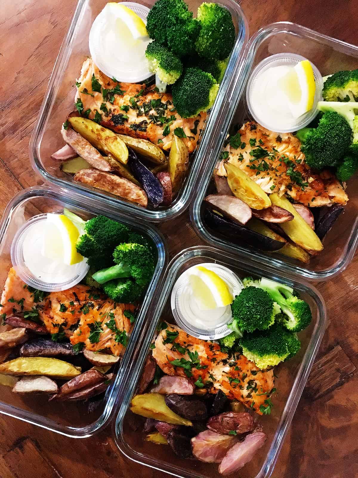 Pan seared salmon with broccoli, fingerling potatoes and garlic aioli with a slice of lemon in glass food prep containers on a brown wood table