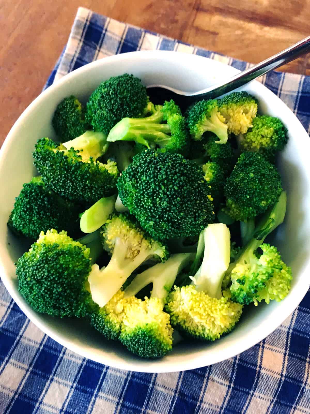 Steamed broccoli in a white bowl on a blue and tan plaid napkin.