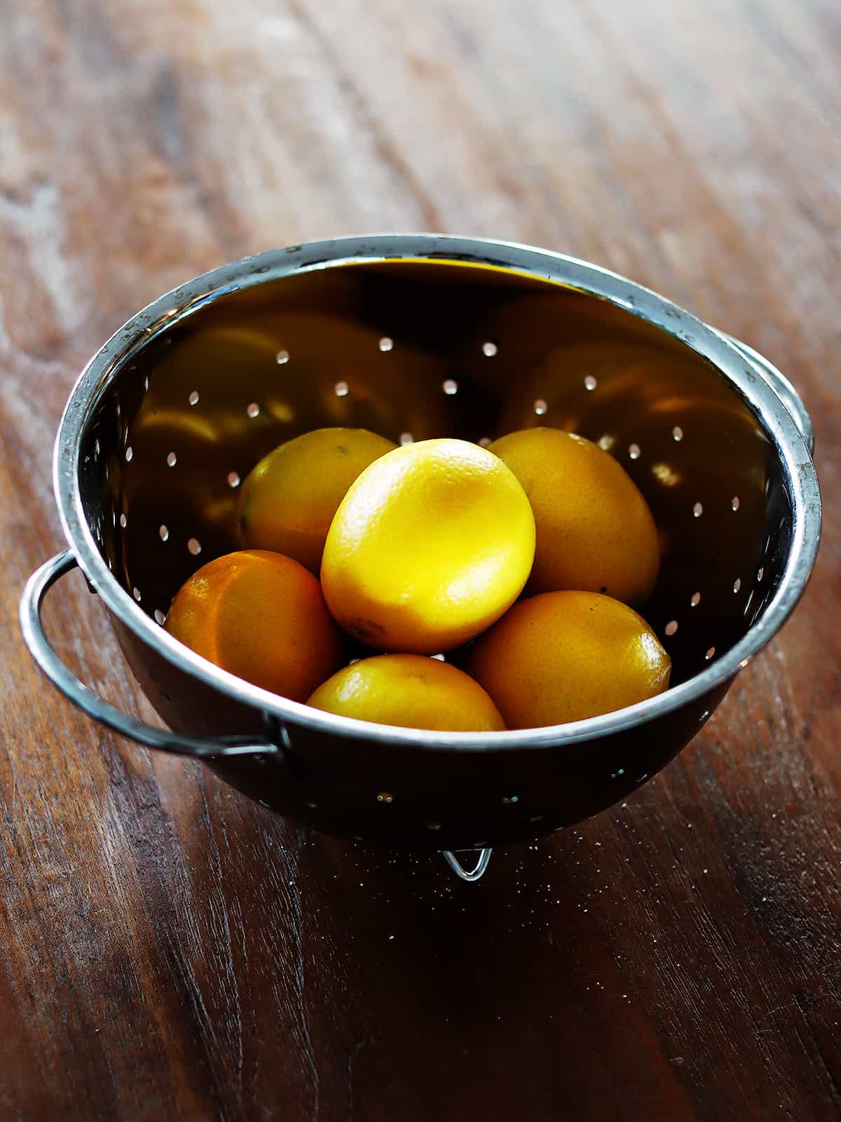 Fresh Meyer lemons in a small stainless steel colander on a brown wood table.