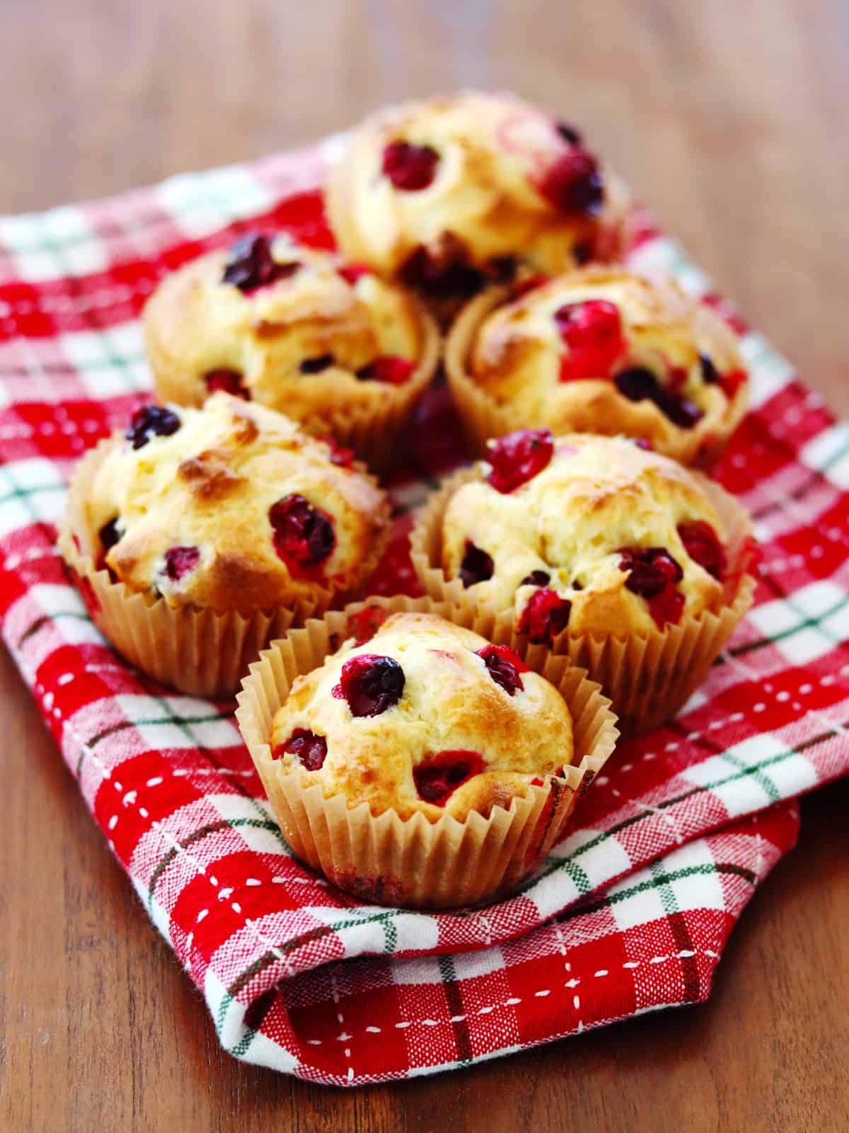 Fresh cranberry orange muffins on a red and white plaid napkin atop a brown wood table.