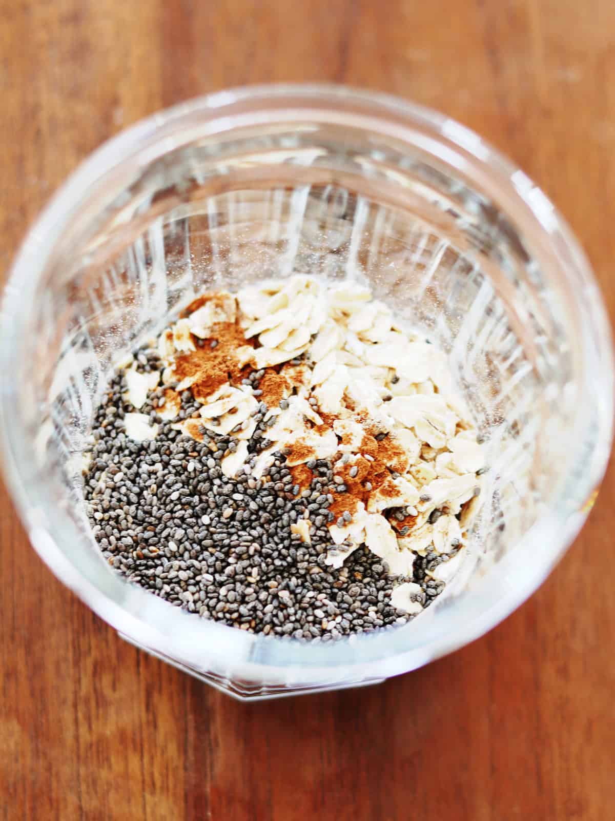 Rolled oats, chia seeds and spices in a glass working jar on a brown wood table.