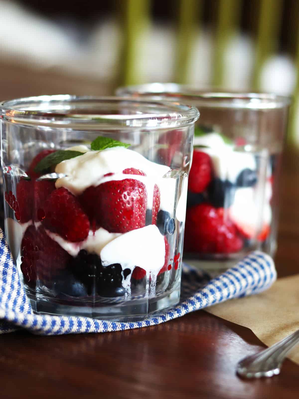 Two berry and yogurt parfaits in glass working jars on a brown wood table.