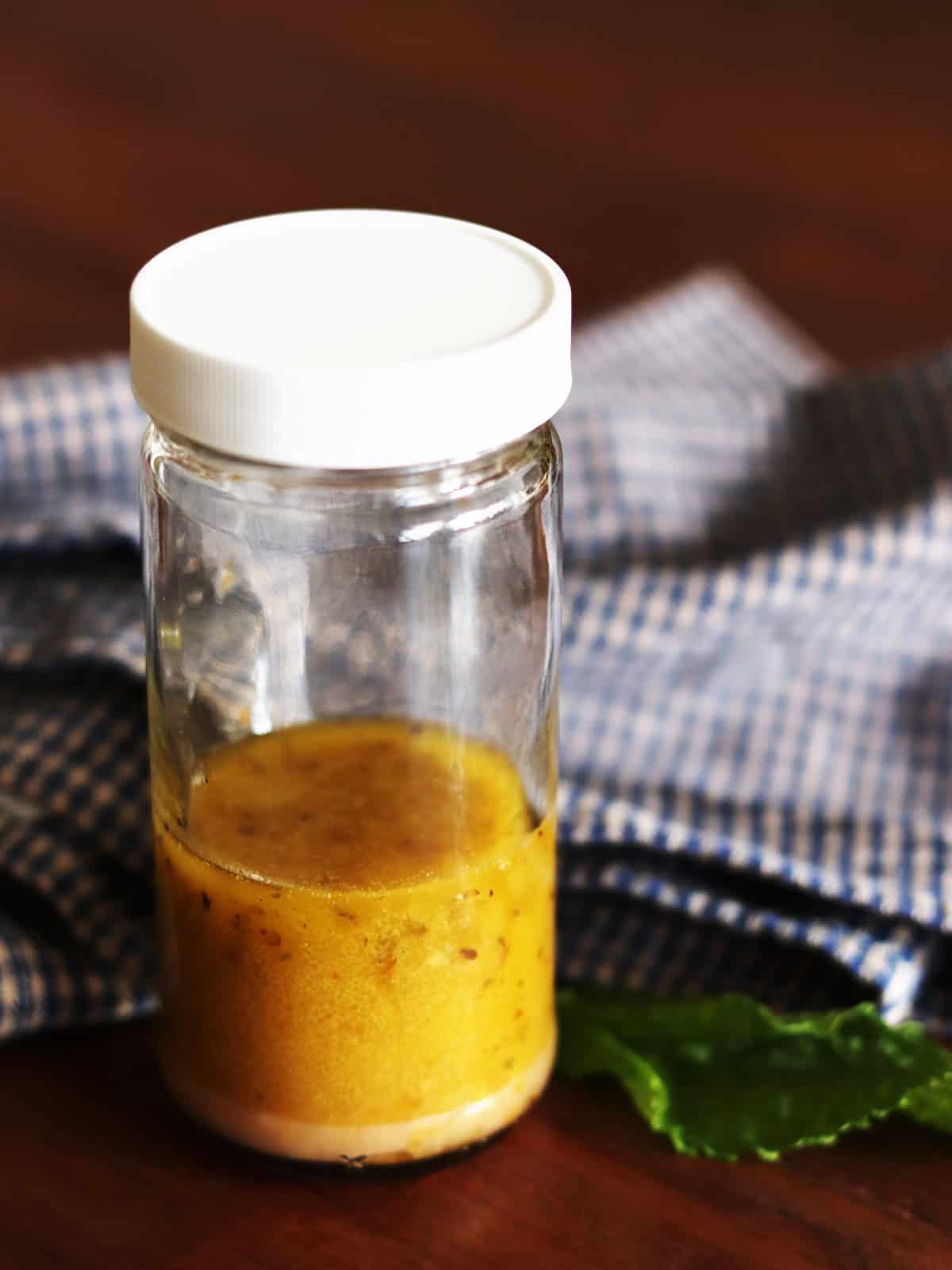 Thin glass bottle of Greek salad dressing with a blue and tan plaid napkin on a brown wood table.