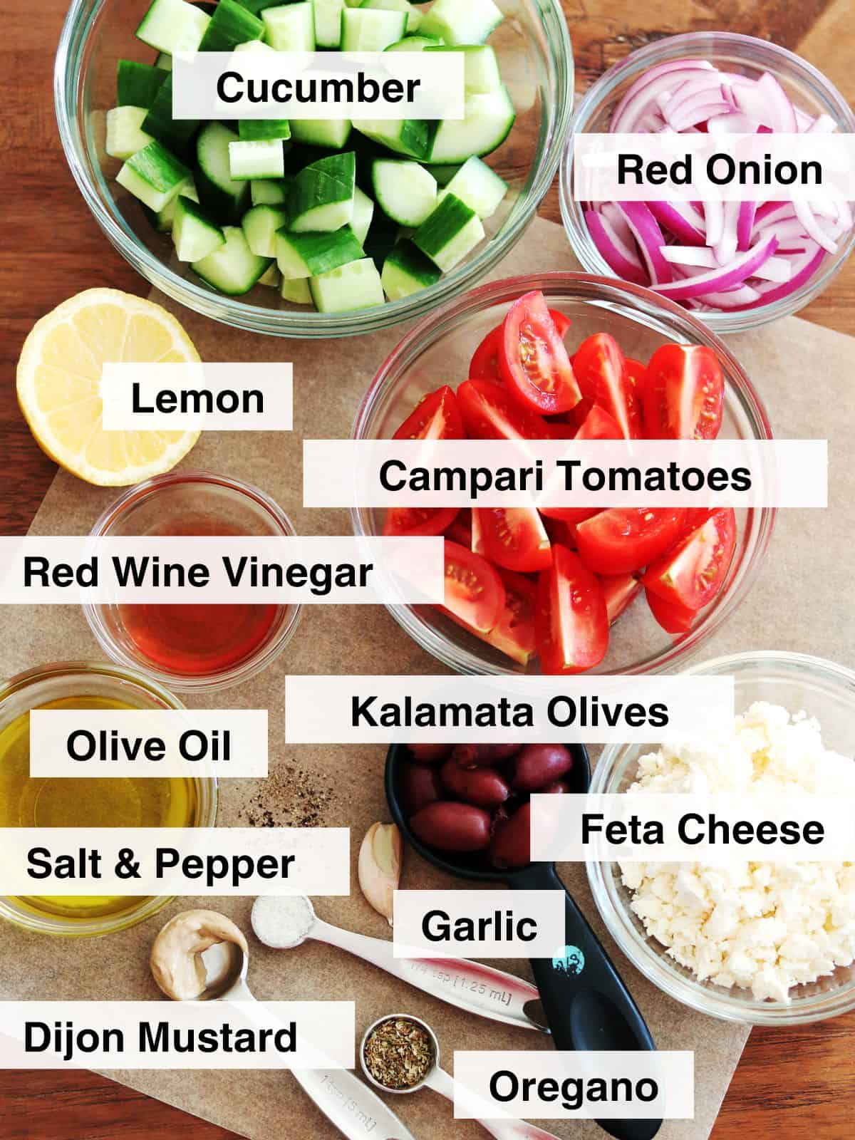 Greek salad ingredients in glass food prep bowls on a brown wood table.