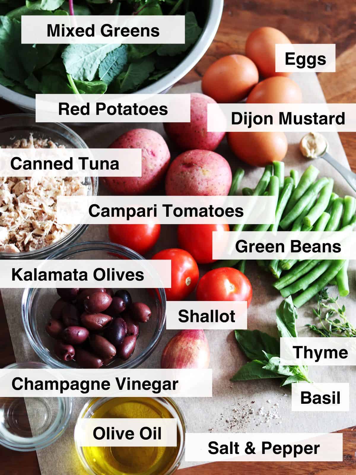 Fresh ingredients for Niçoise salad arranged on a brown wood table lined with parchment paper.