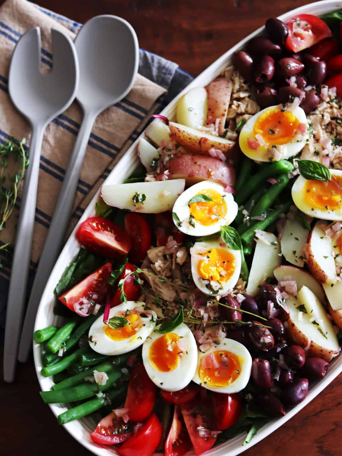 Homemade Niçoise salad in a ceramic beige serving platter with grey serving utensils.