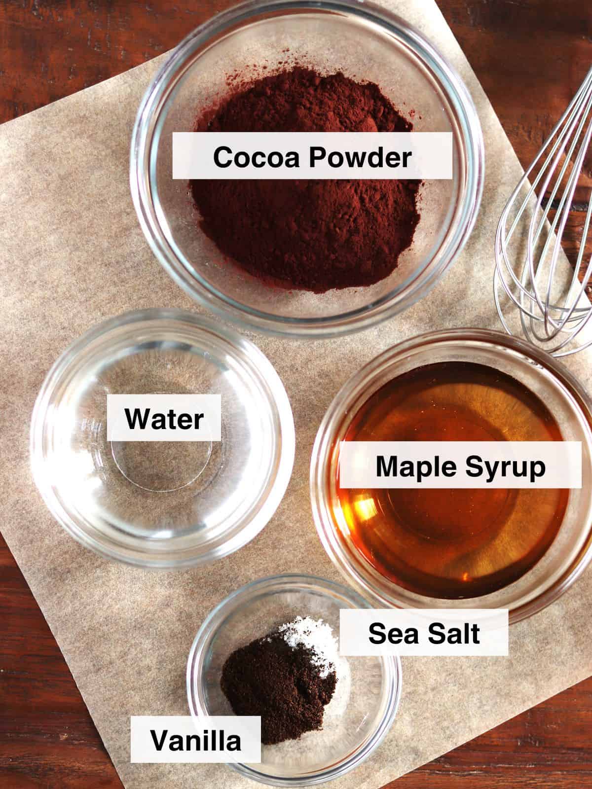 Chocolate sauce ingredients in glass meal prep bowls on a parchment-paper lined wood table.