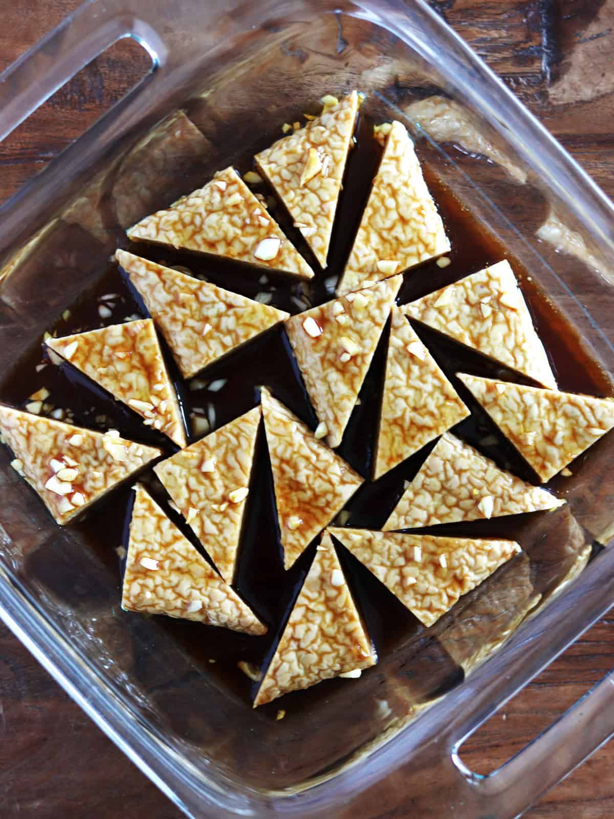 Tempeh triangles marinating in a tamari maple sauce inside a glass baking dish.