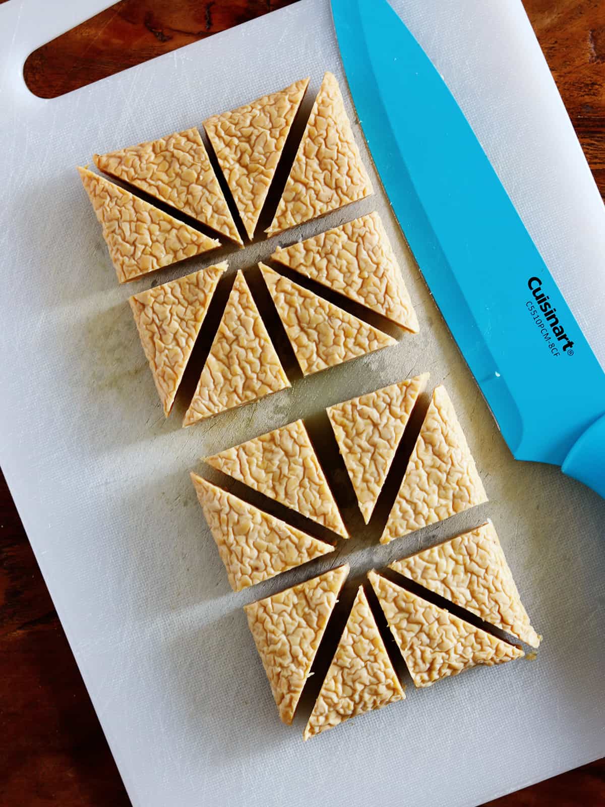 Tempeh cut into triangles on a white silicone cutting board with a light blue ceramic chef's knife.