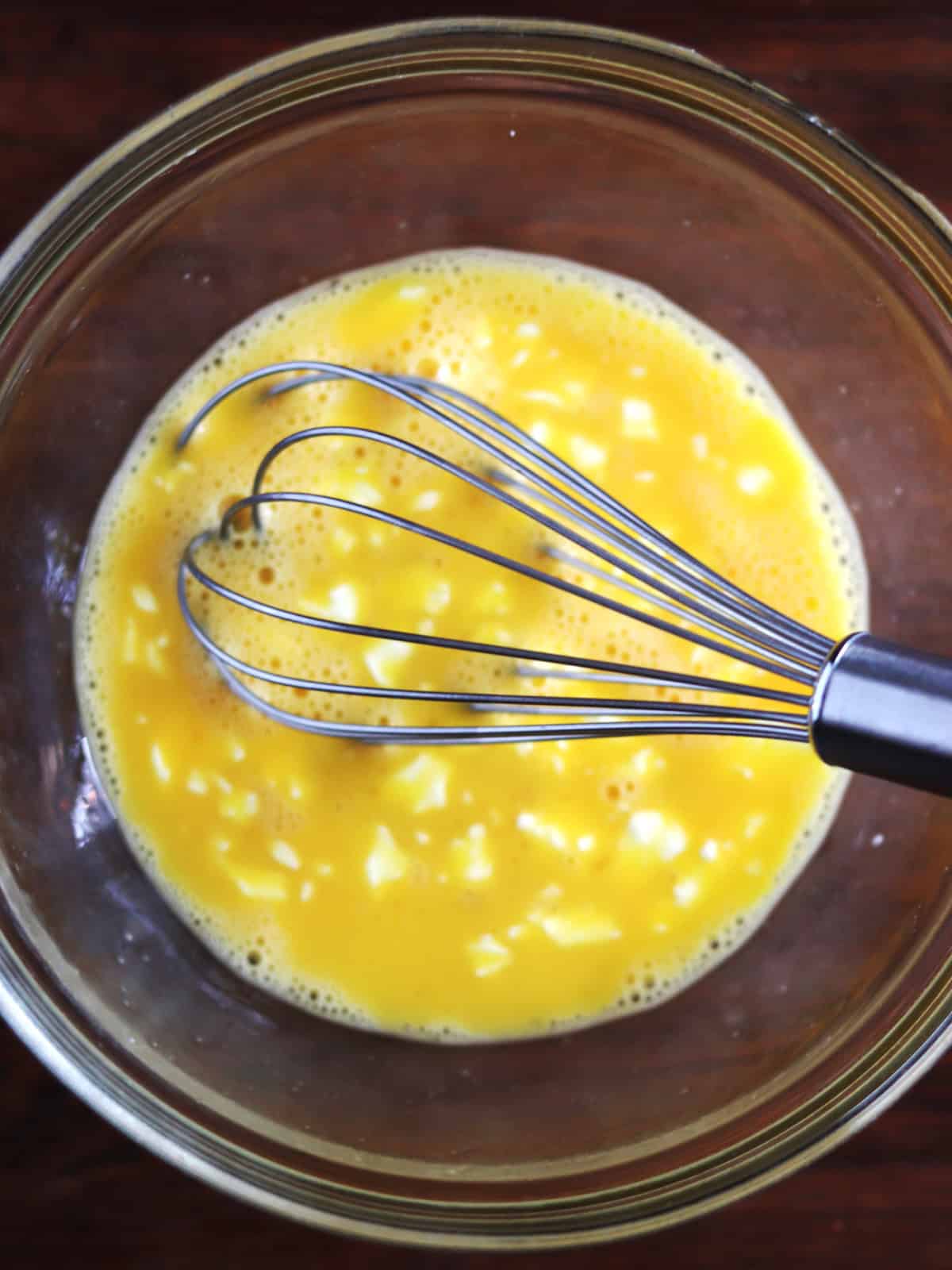 Whisked eggs with chunks of cream cheese in a glass bowl.