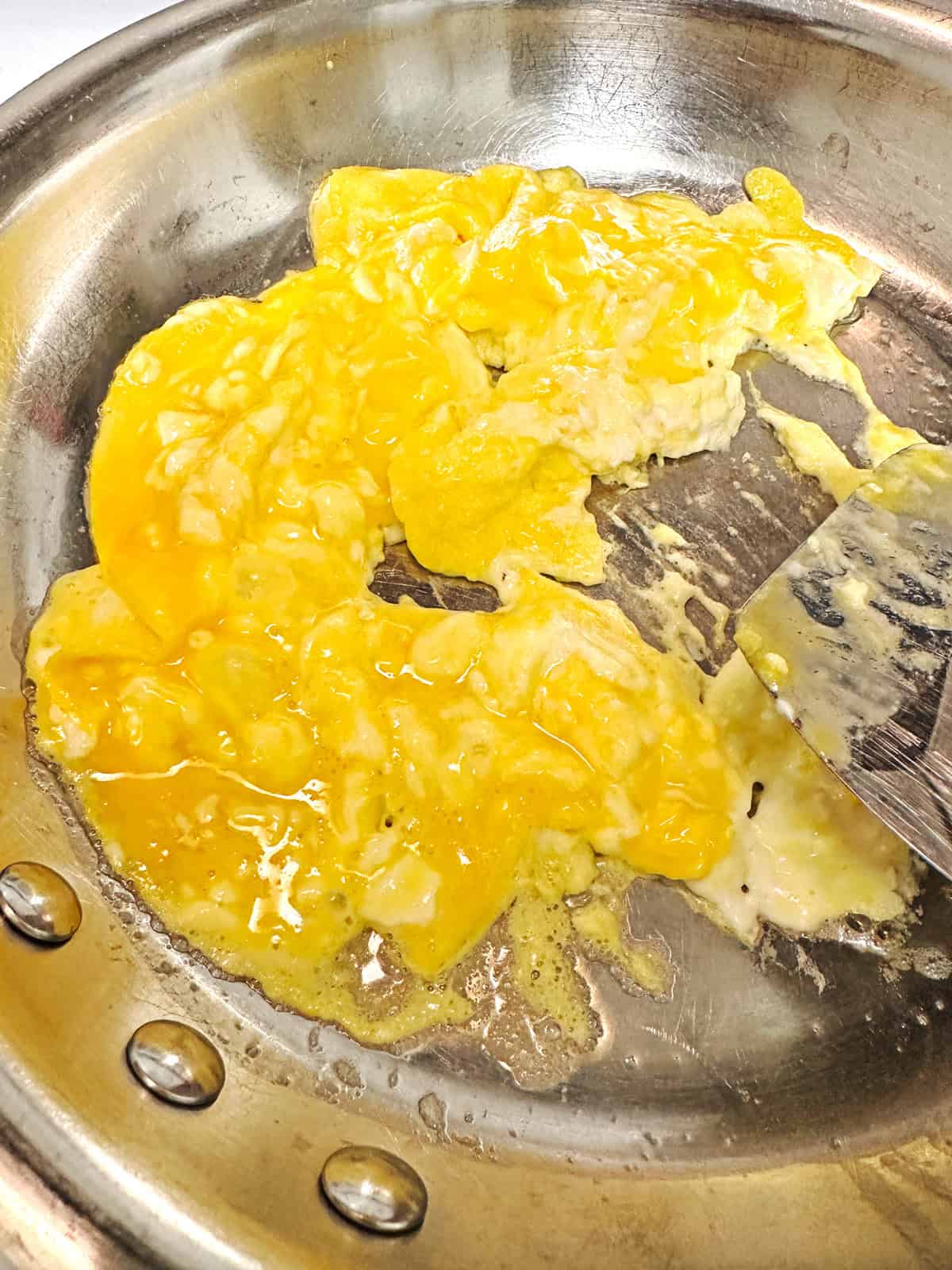 Whisked eggs and cream cheese in a stainless steel skillet with a metal spatula.