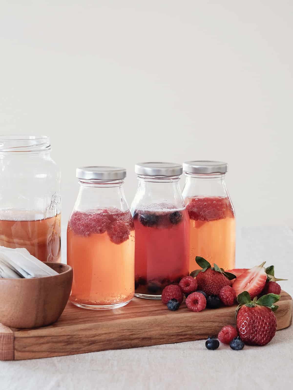 Glass jars of kombucha with fresh raspberries, blueberries and strawberries added to and placed around the jars.