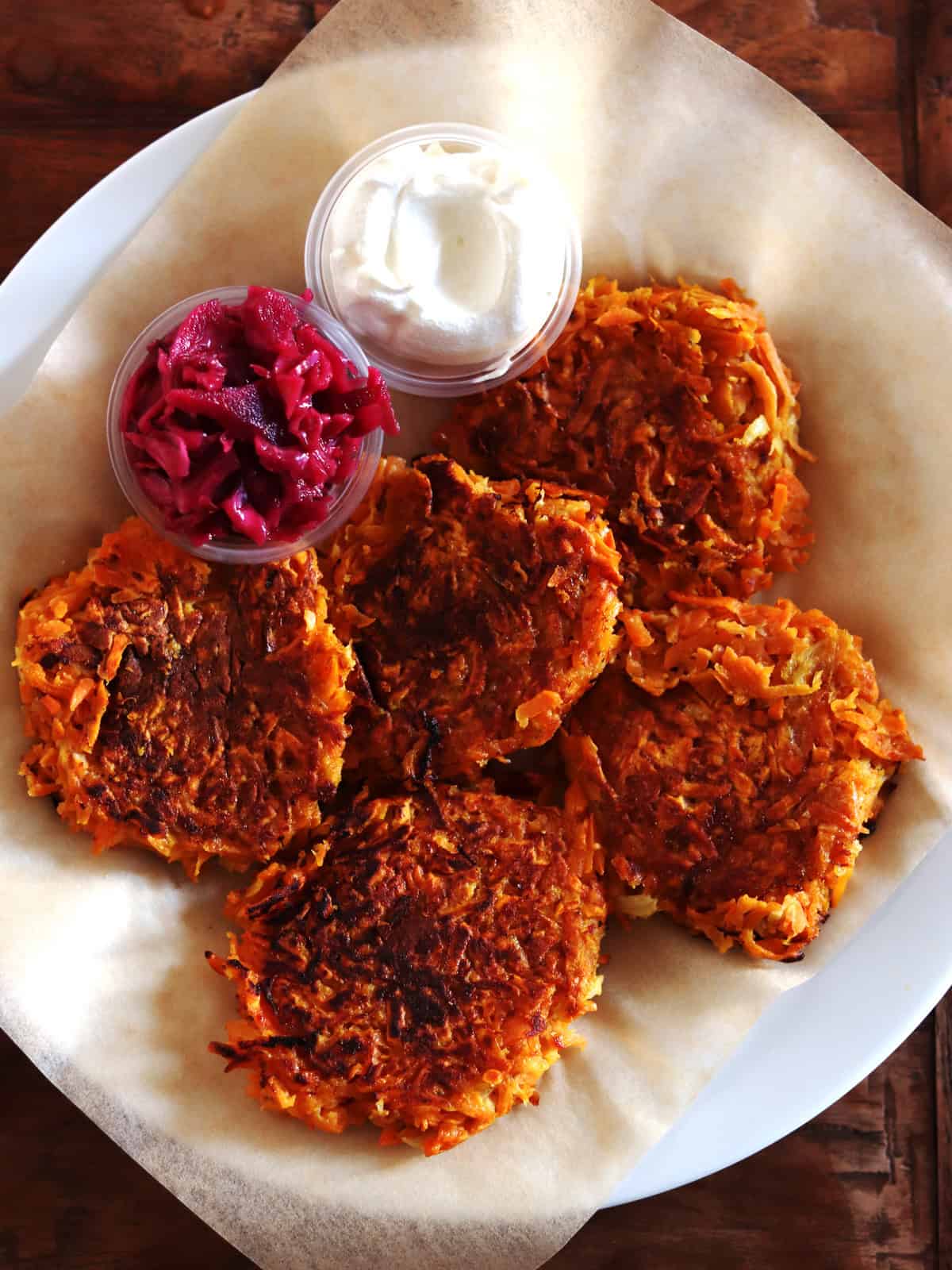 Sweet potato latkes with sides of beet kraut and sour cream on a parchment paper lined plate.