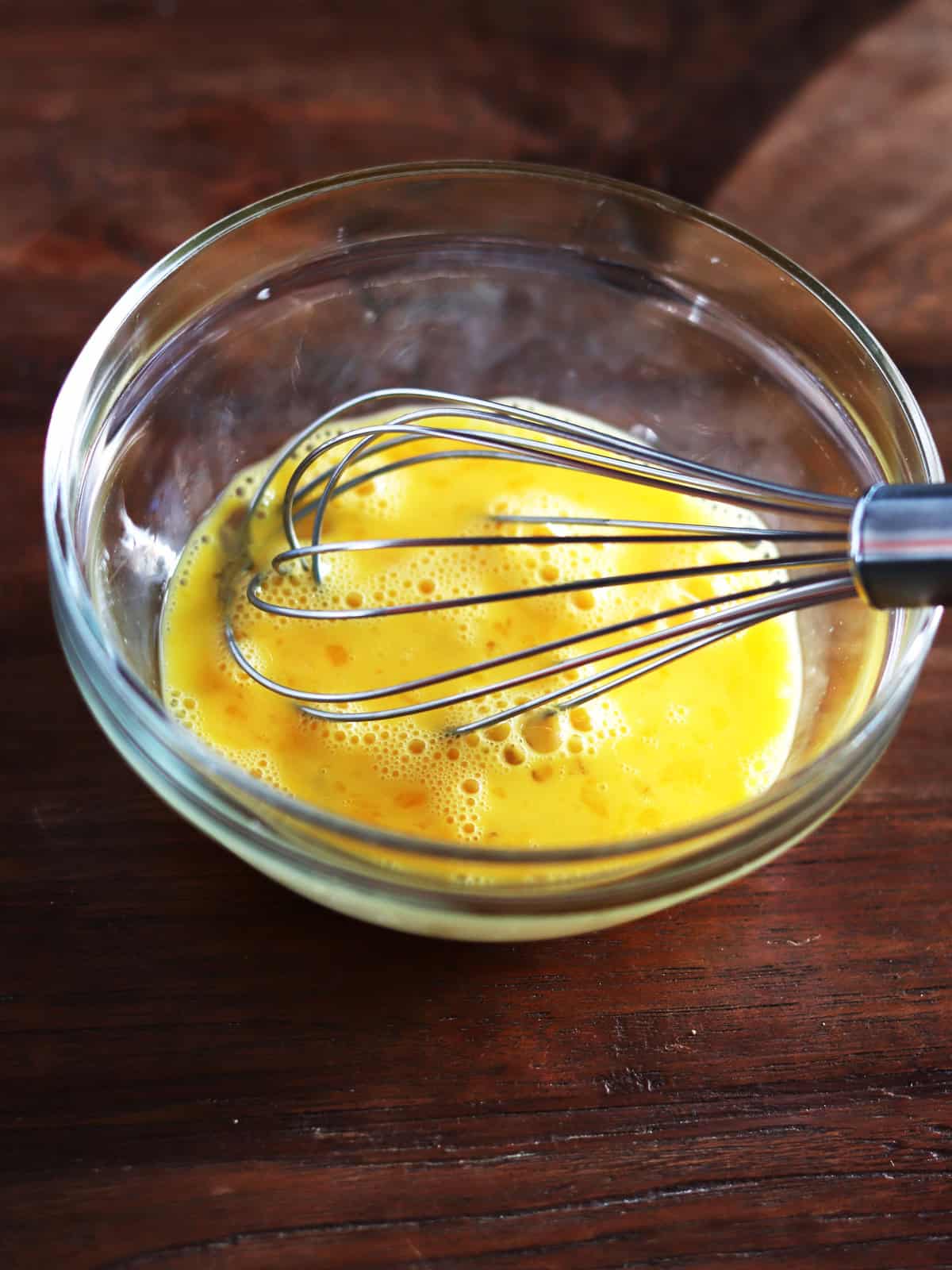 Whisked eggs in a glass mixing bowl with a stainless steel whisk on a brown wood table.
